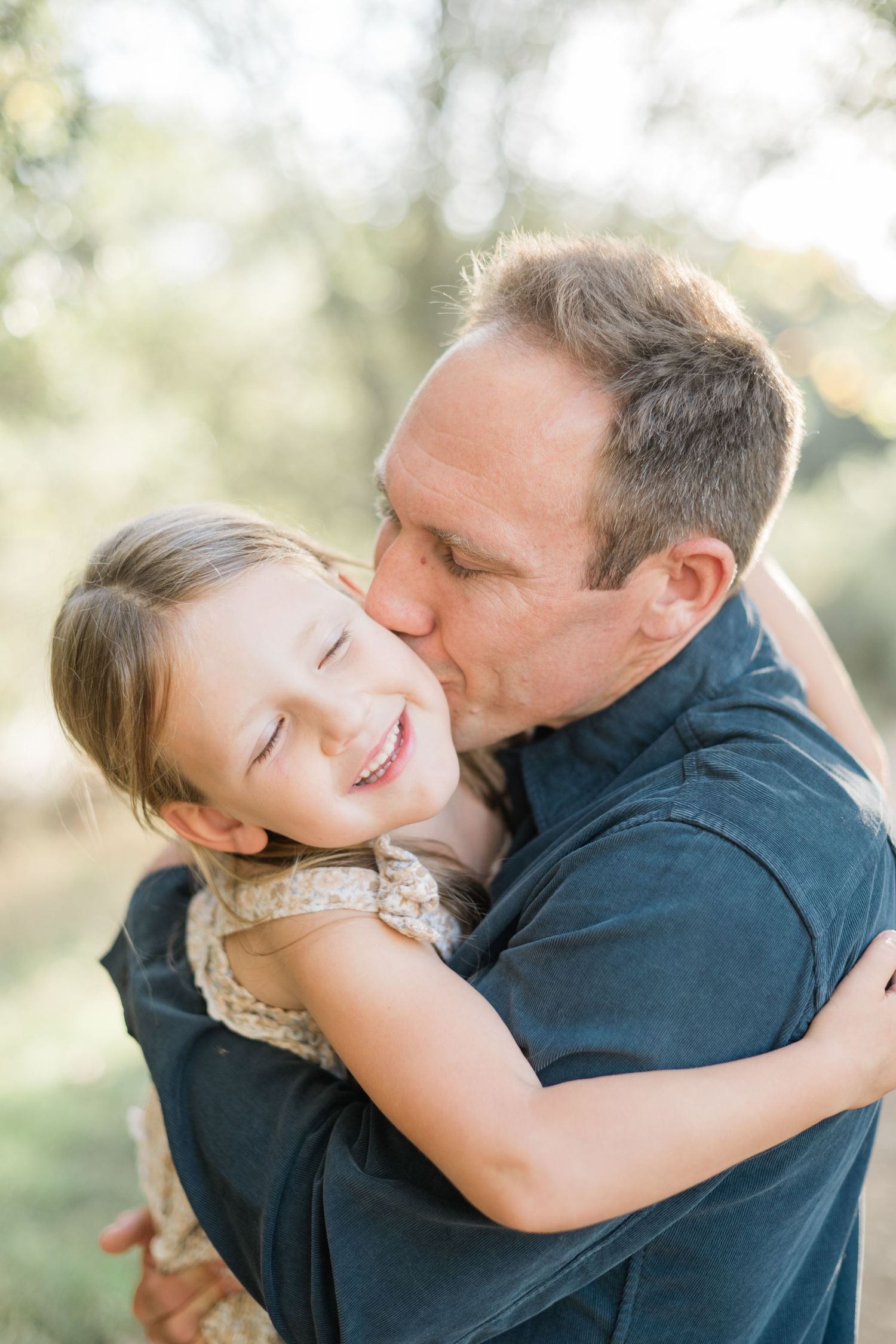 A father kisses his smiling young daughter on the cheek while hugging her outdoors in soft natural light.