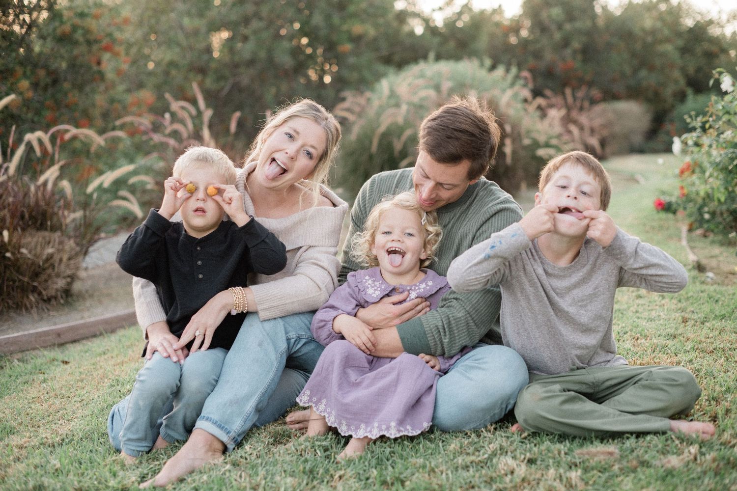 Happy family of five sitting on grass, parents and three children laughing and making funny faces outdoors.