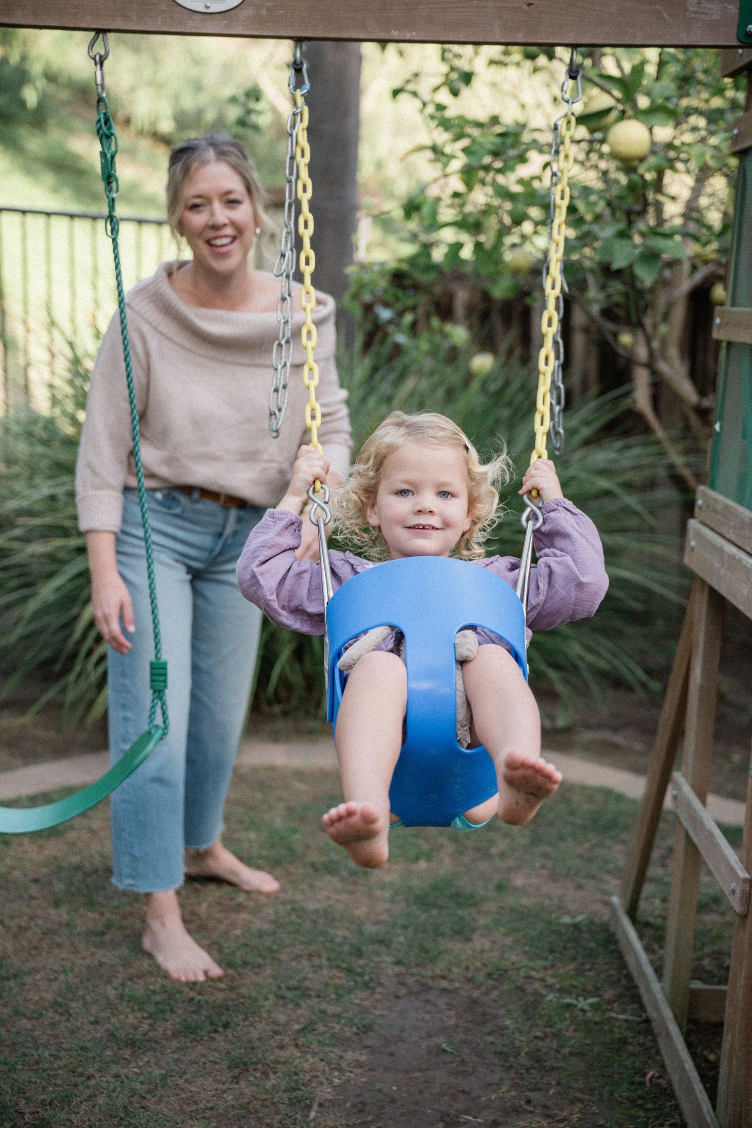 A smiling mother pushes her happy toddler girl on a blue bucket swing in a backyard playground.