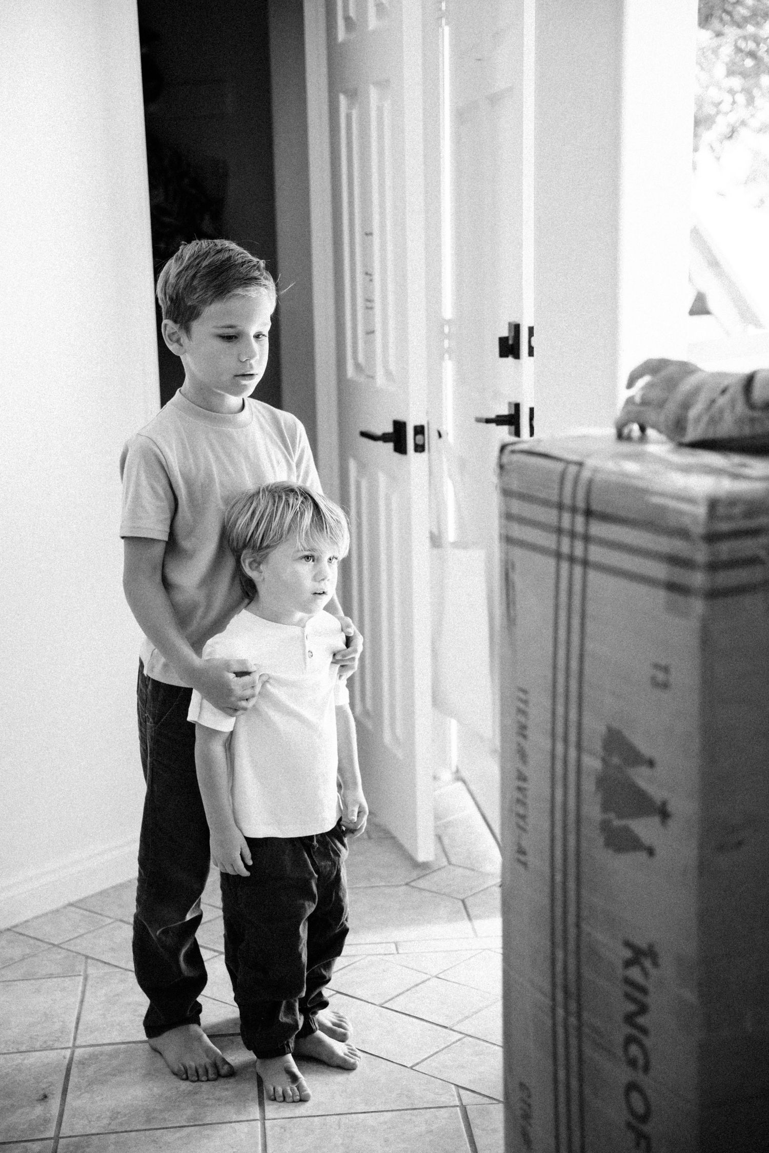 Two young boys standing together near a doorway with moving boxes in black and white photo.
