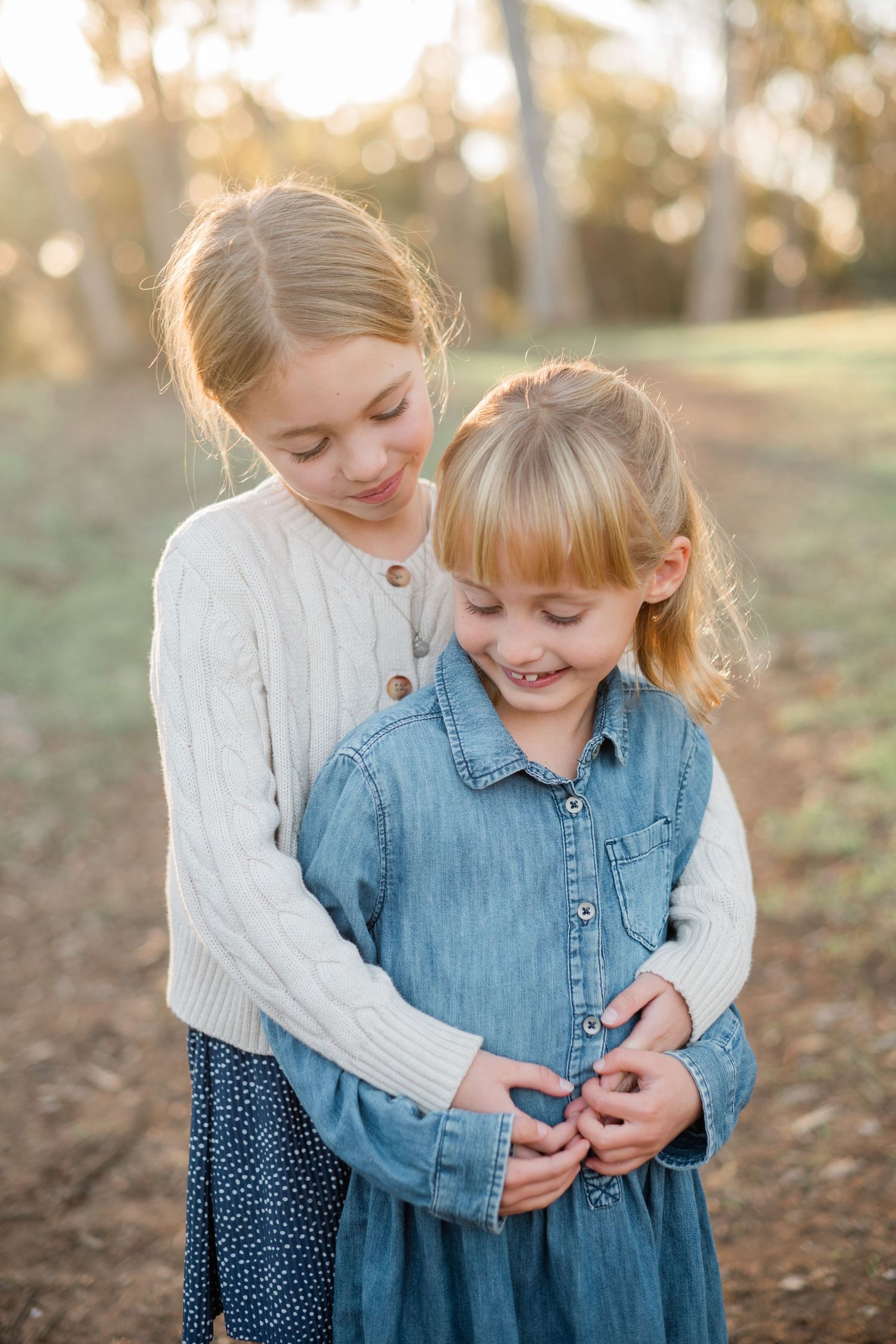 Two young blonde sisters share a sweet embrace outdoors at golden hour, older girl hugging younger in denim dress.