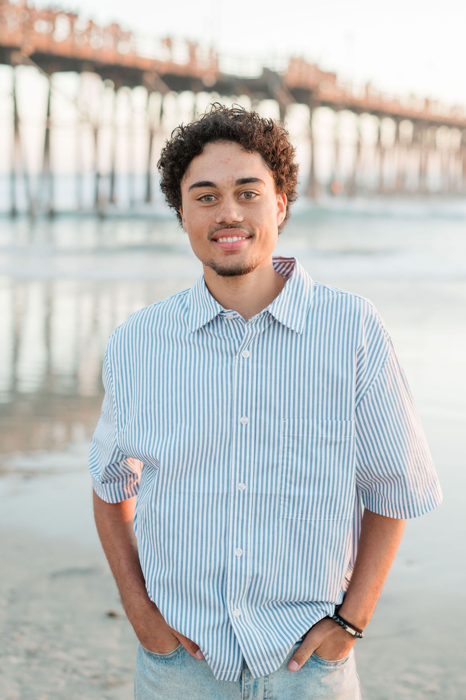 Smiling young man with curly hair in a striped shirt standing on the beach with a pier in the background.