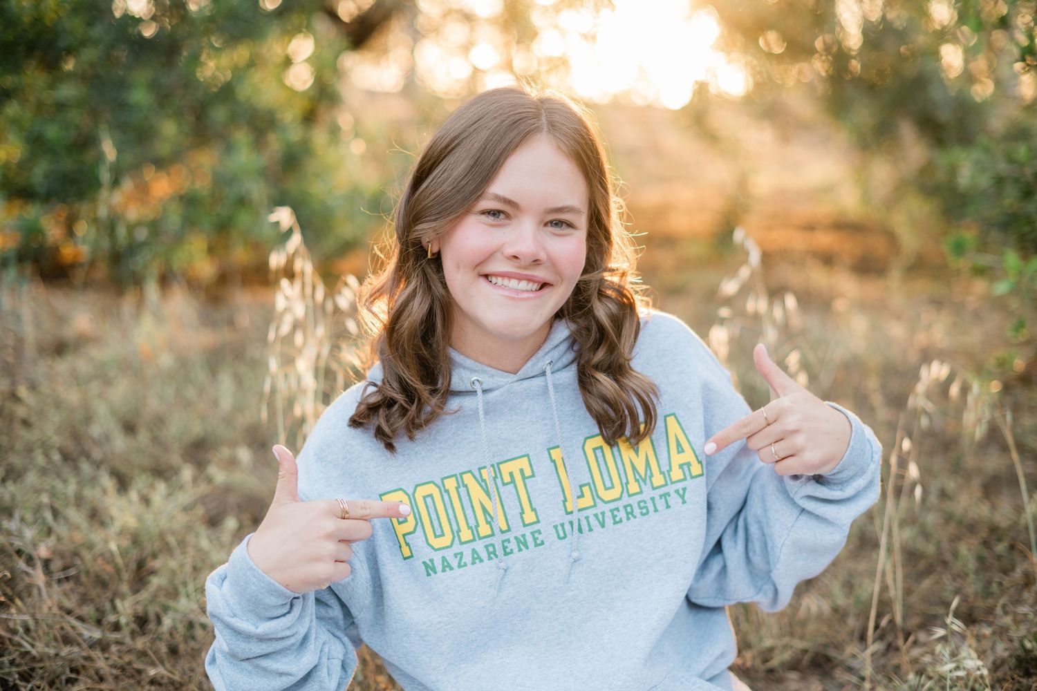 Smiling young woman proudly pointing to her Point Loma Nazarene University hoodie outdoors.