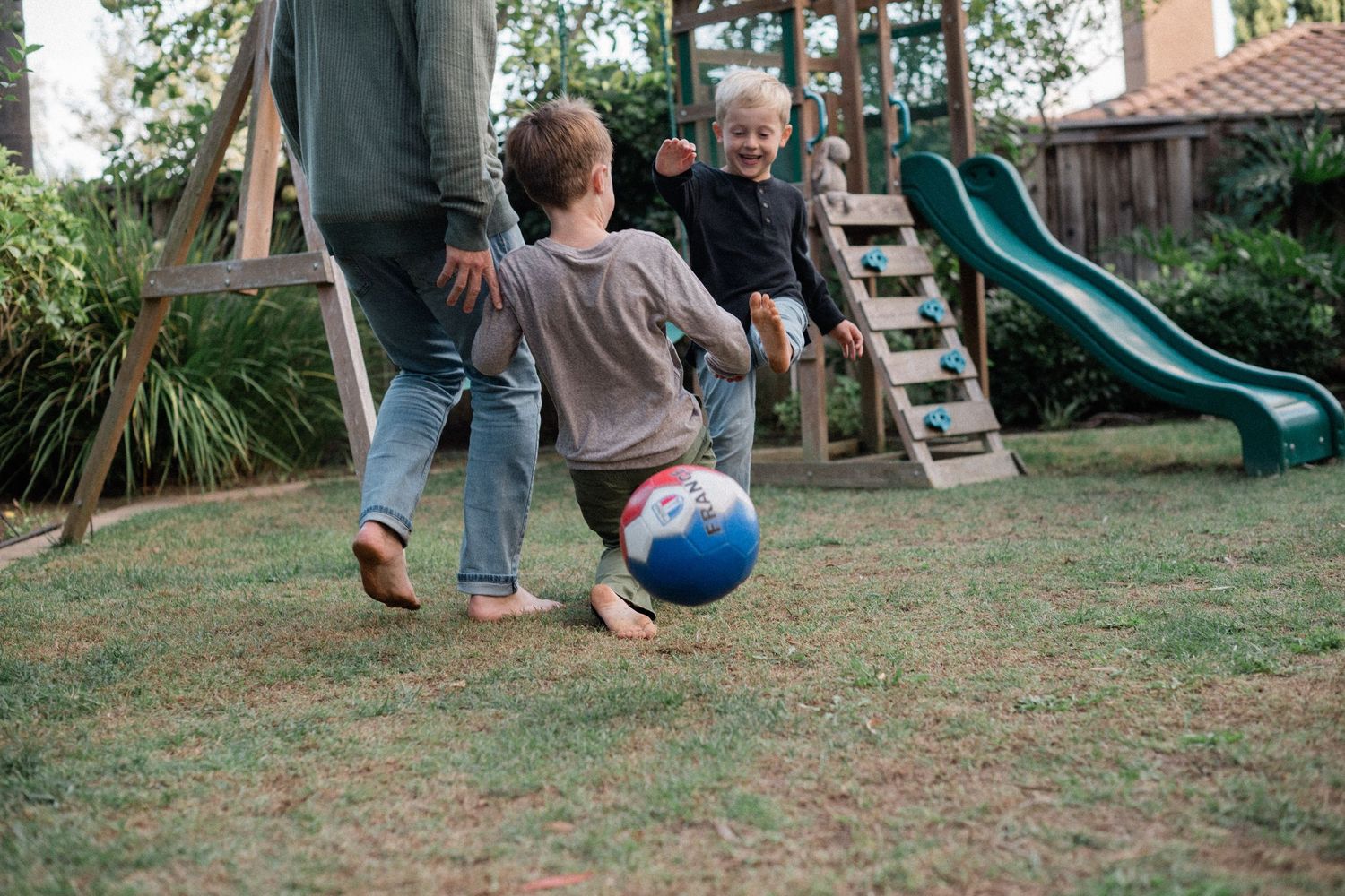 Two children playing soccer with an adult in a backyard with a green slide and playground equipment.