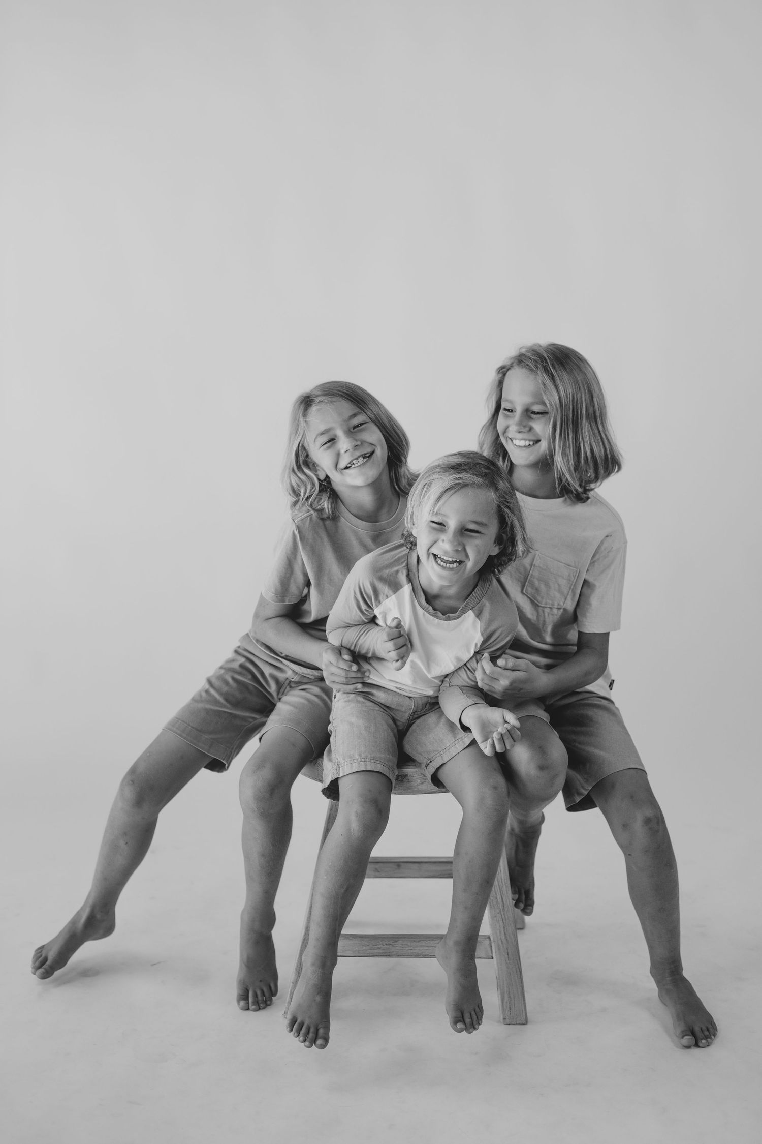 Black and white studio portrait of three smiling children sitting together on a wooden stool.