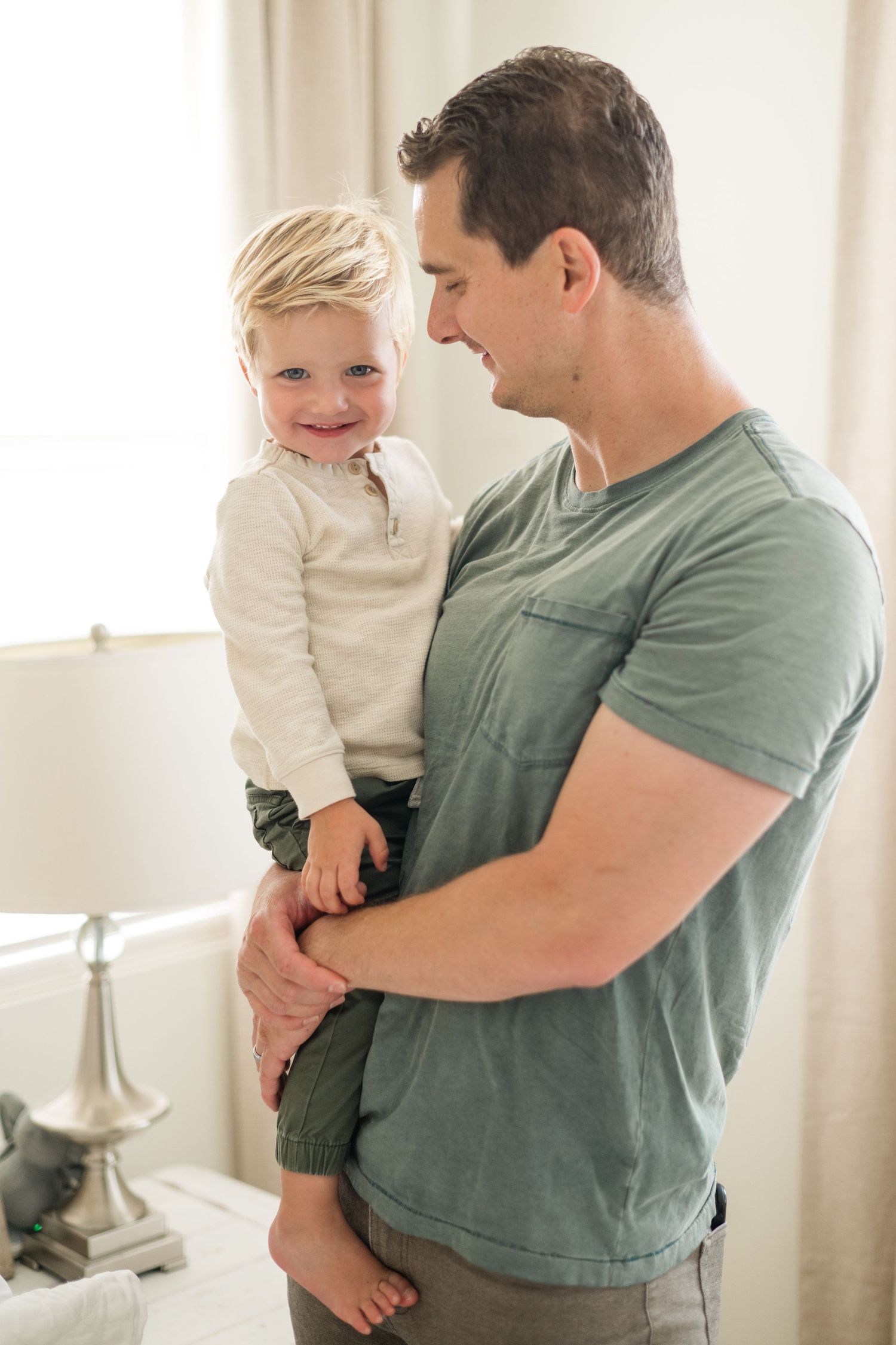 A smiling toddler boy held by his father in a green t-shirt in a bright bedroom.