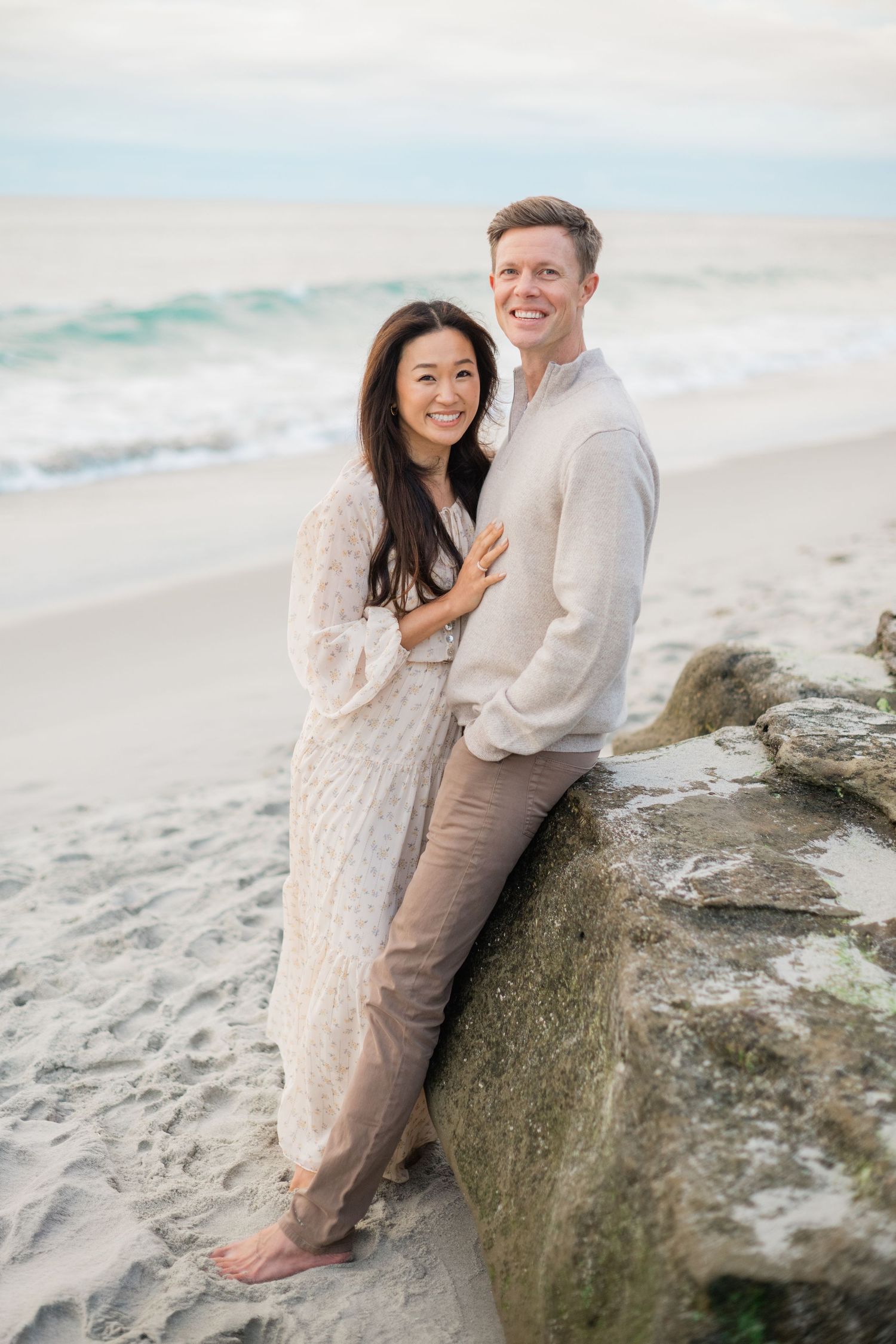 Smiling couple posing together on a sandy beach near rocks with ocean waves in the background.