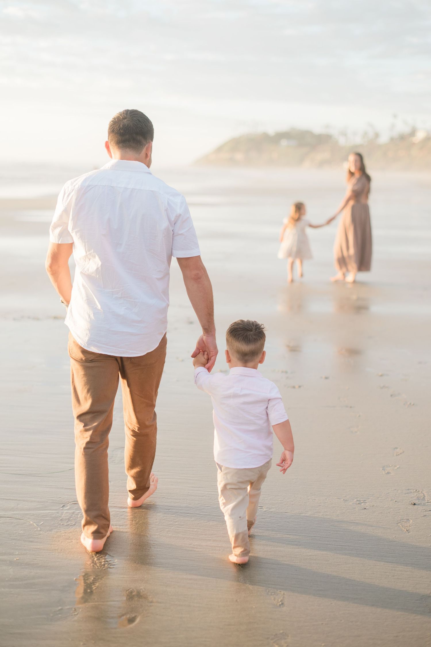 Father and son walk hand-in-hand on a sunlit beach toward mother and daughter in a heartwarming family moment.