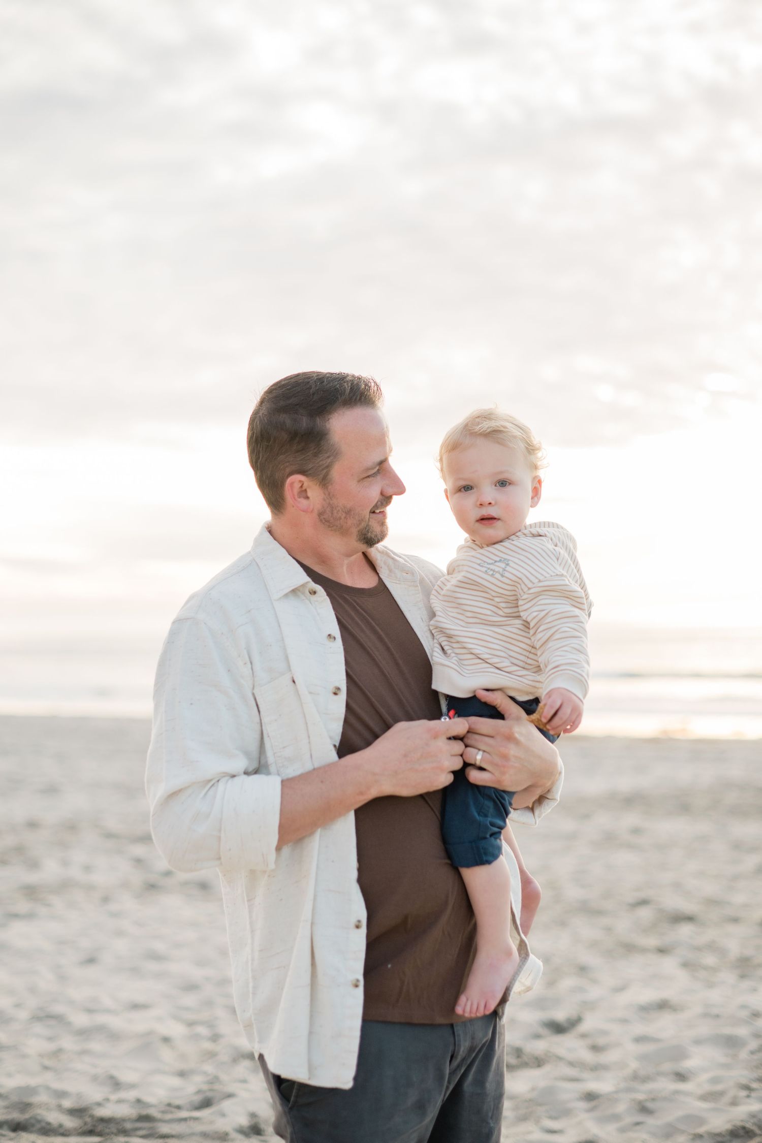 A father holds his toddler on a sandy beach, both smiling, with a cloudy sky in the background.