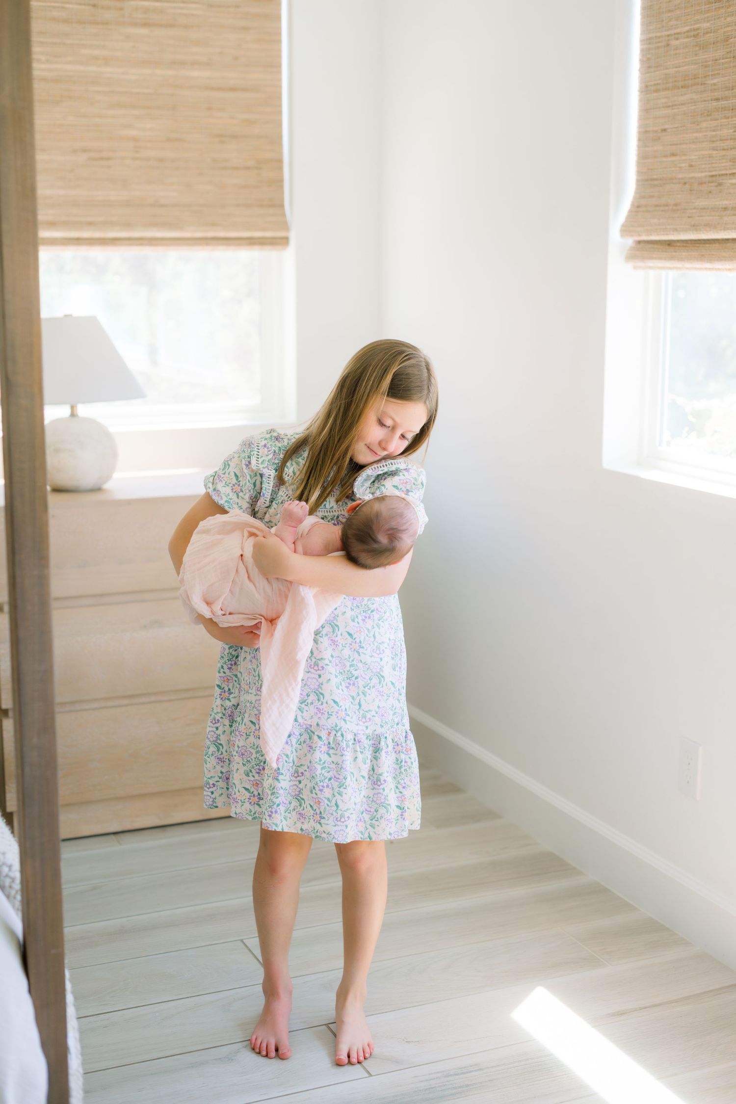 A young girl in a floral dress tenderly holds a newborn baby wrapped in a pink blanket in a bright, airy room.