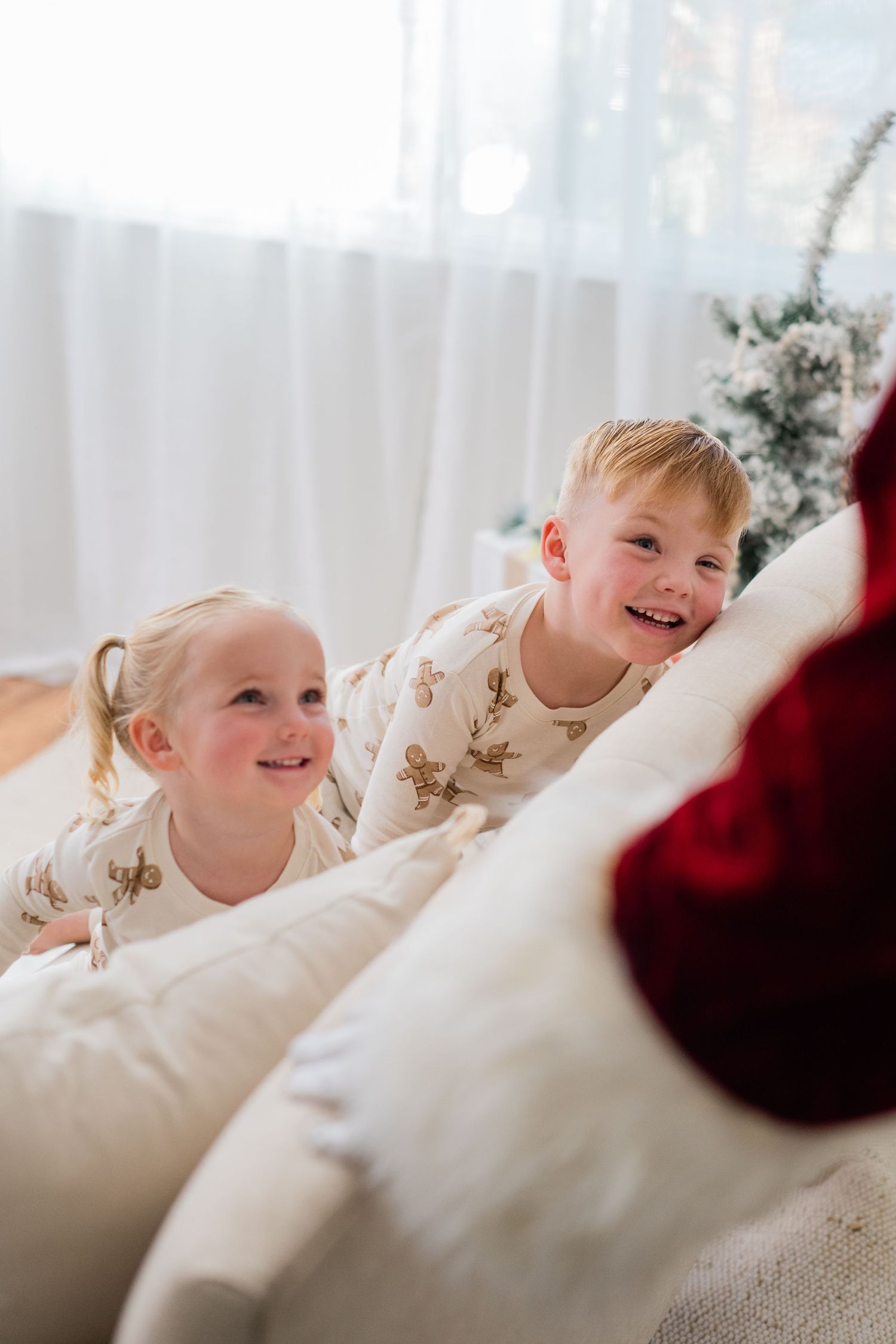 Two smiling children in festive pajamas sit on a white couch near a Christmas tree, excited for the holiday season.