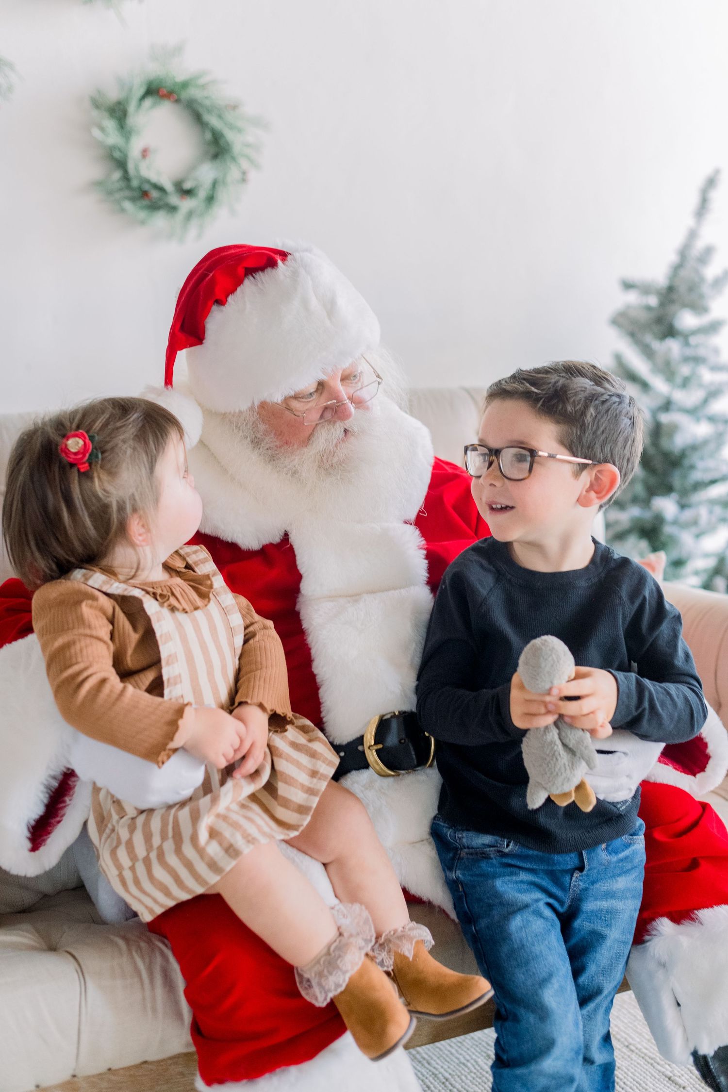 Two young children sit with Santa Claus on a couch, smiling and interacting near a decorated Christmas tree.