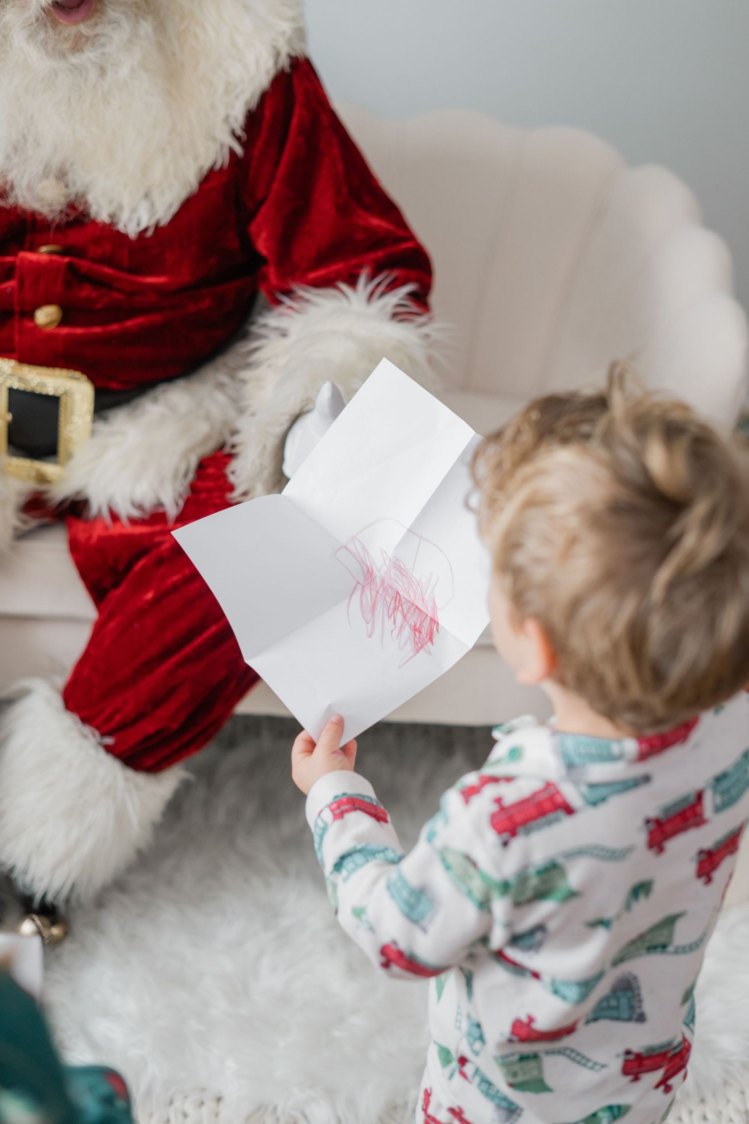A toddler in Christmas pajamas shows Santa Claus a hand-drawn picture during a holiday photo session.