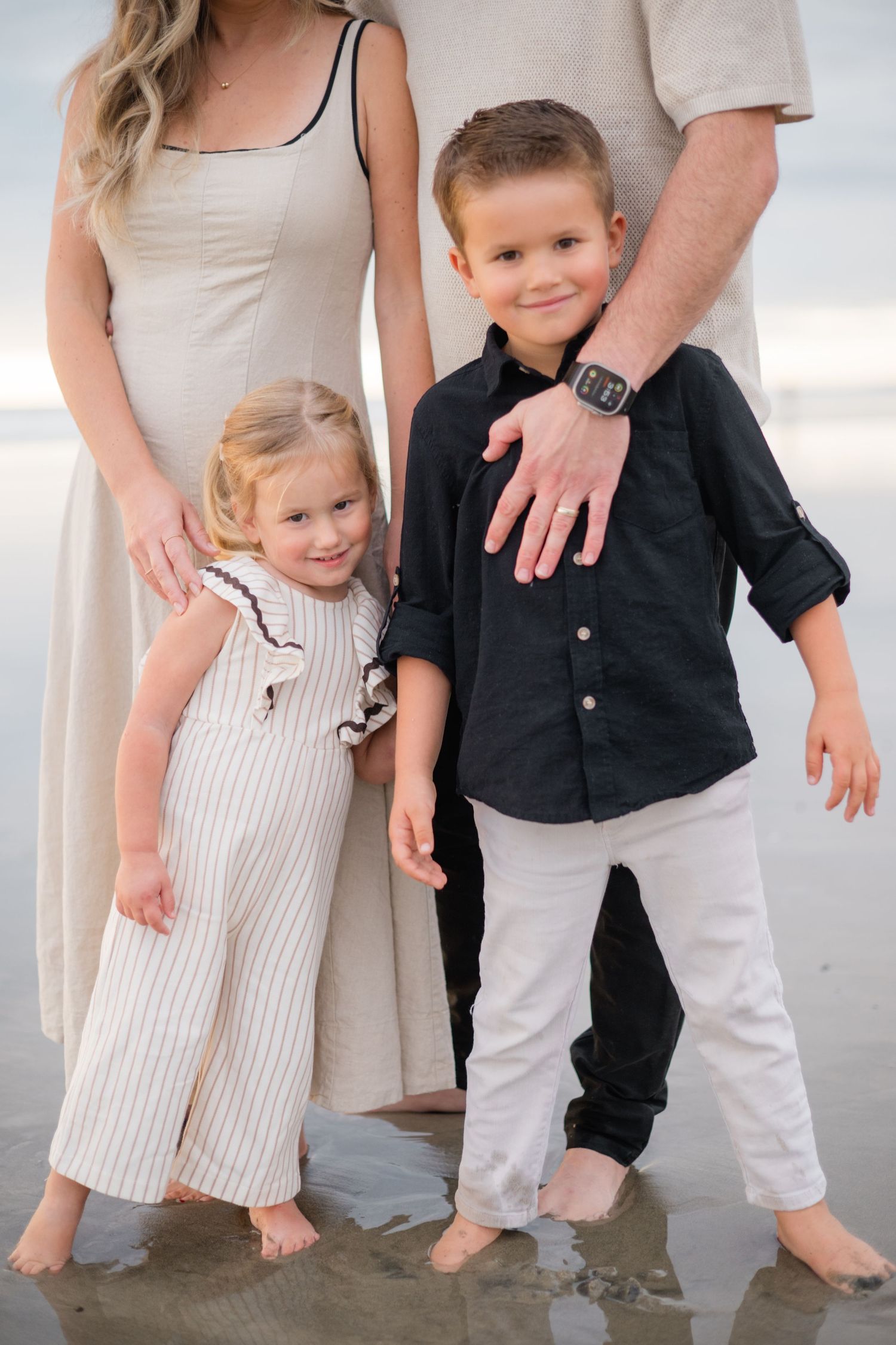 Two young siblings standing with parents on a beach, boy in black shirt and girl in striped romper smiling.