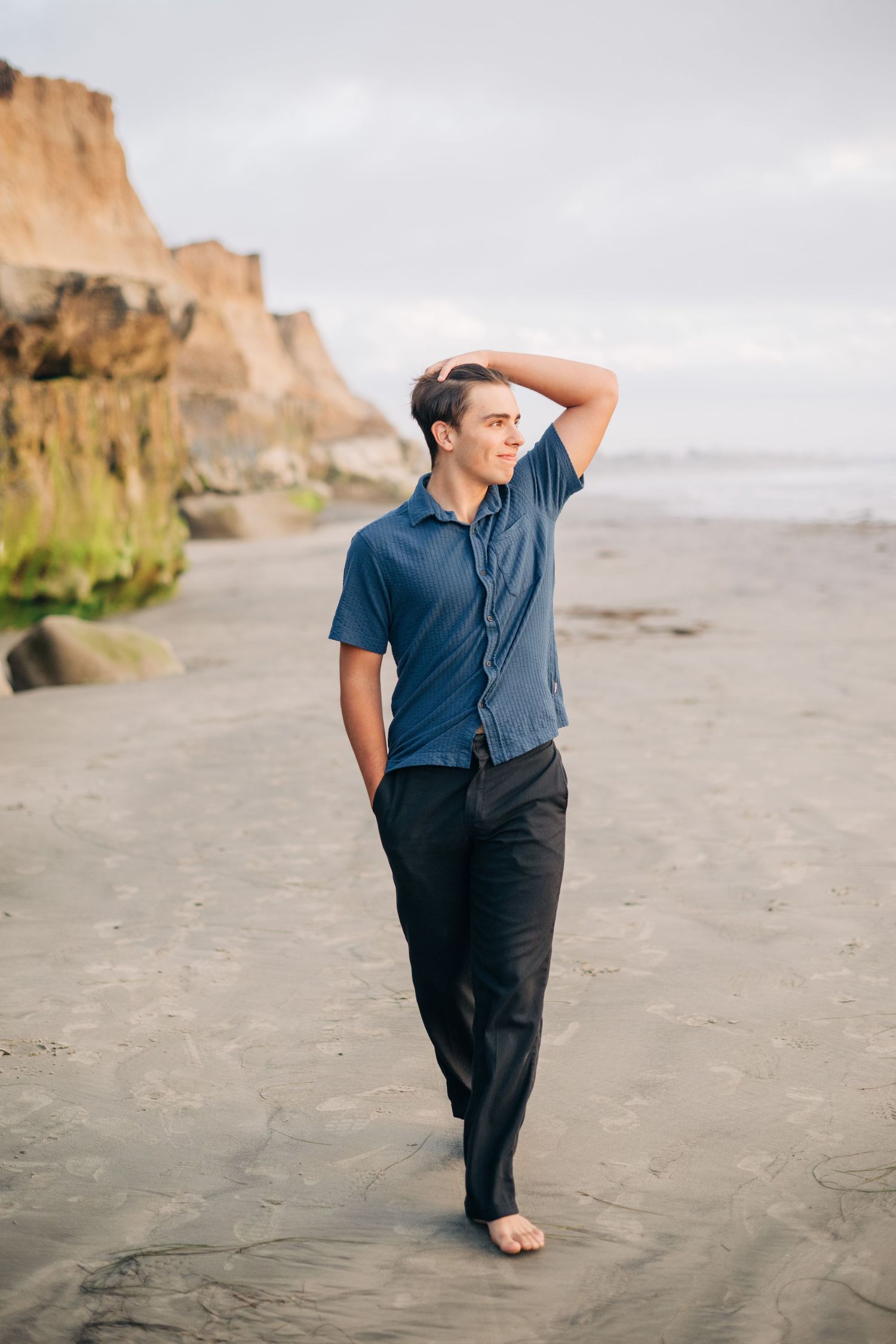Young man in blue shirt and black pants poses barefoot on sandy beach with rocky cliffs in background.