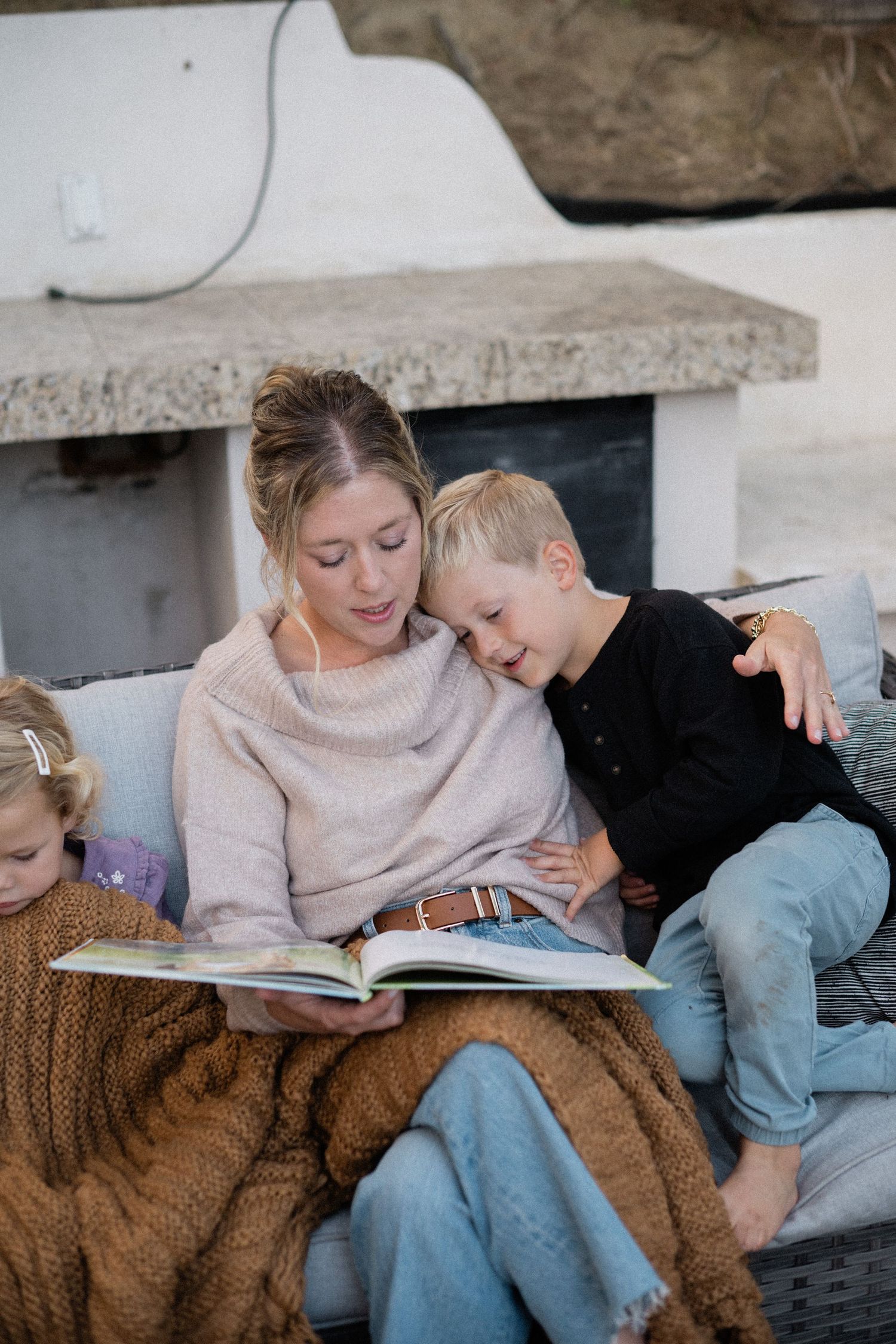 A blonde woman in beige sweater reads a picture book to two young children on an outdoor sofa by fireplace.