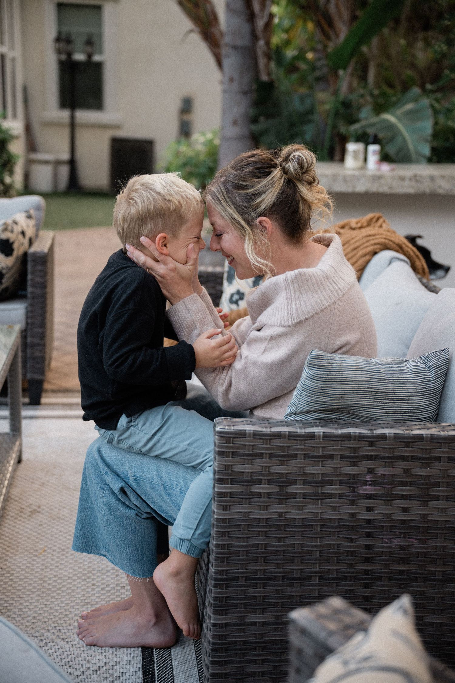 Mother and young blonde son share a tender forehead-to-forehead moment on a wicker patio sofa.