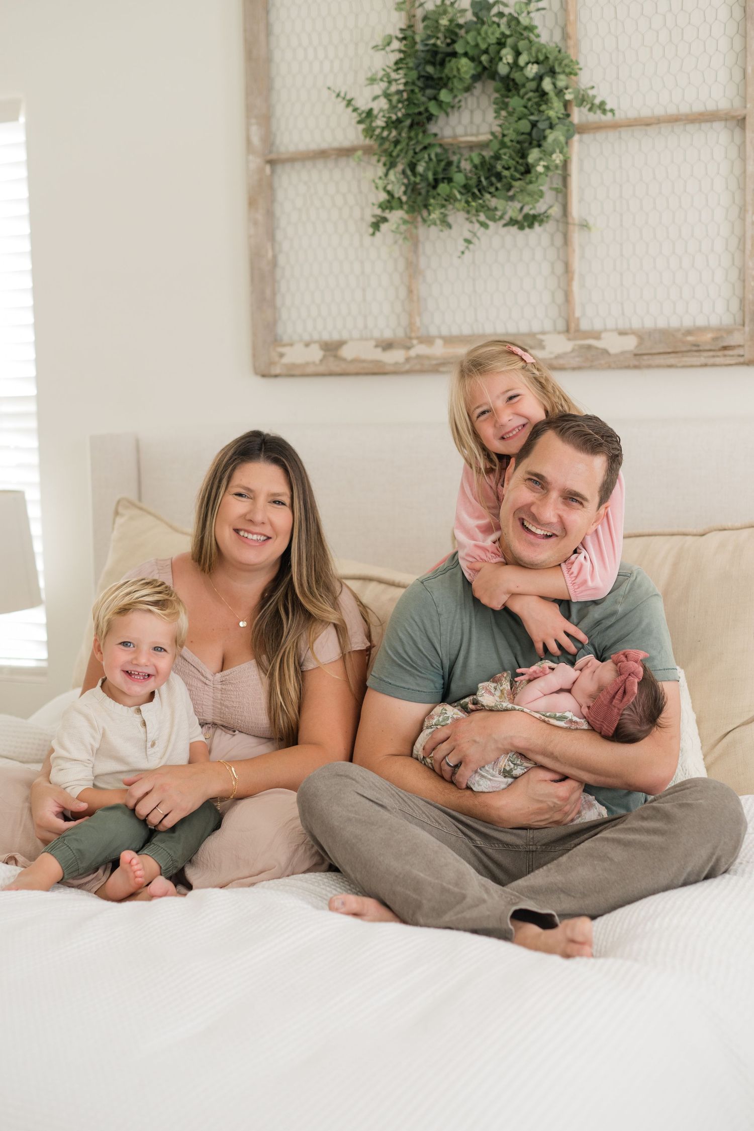 Happy family of five posing on a bed, parents with three children including a newborn, in a bright farmhouse-style bedroom.