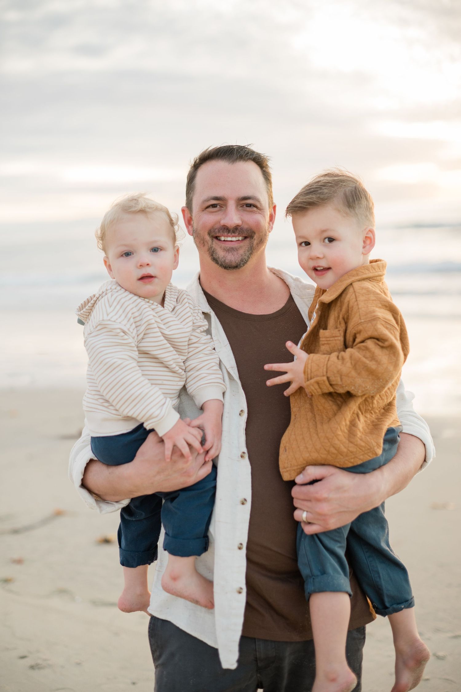 A smiling father holding two young boys on a beach at sunset.