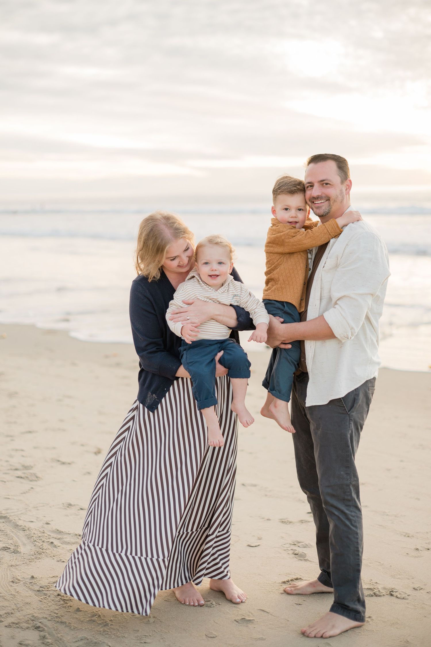 Family of four smiling on beach at sunset, mom in striped skirt holding toddler, dad carrying young boy.