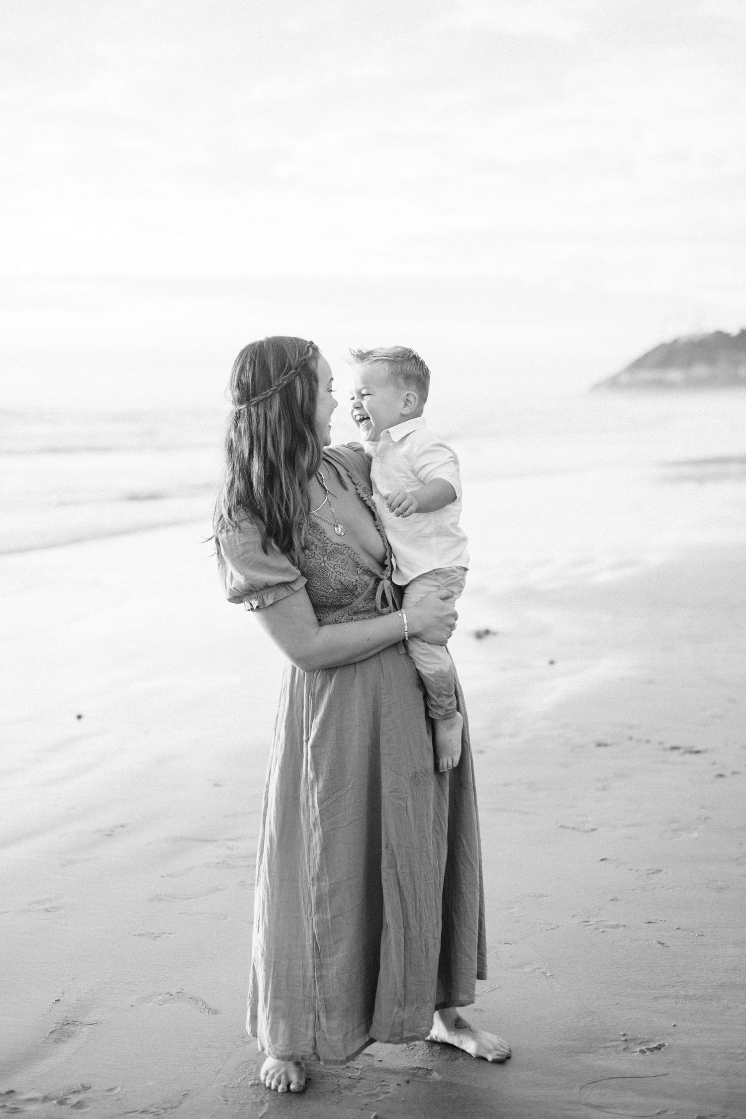 Black and white photo of a mother holding her young son on a beach, sharing a joyful moment by the ocean.