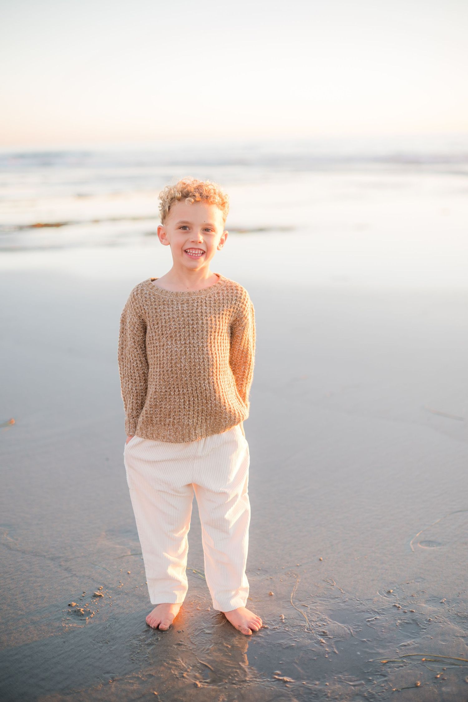 A smiling young boy in a tan knit sweater and white pants stands barefoot on a sandy beach at sunset.