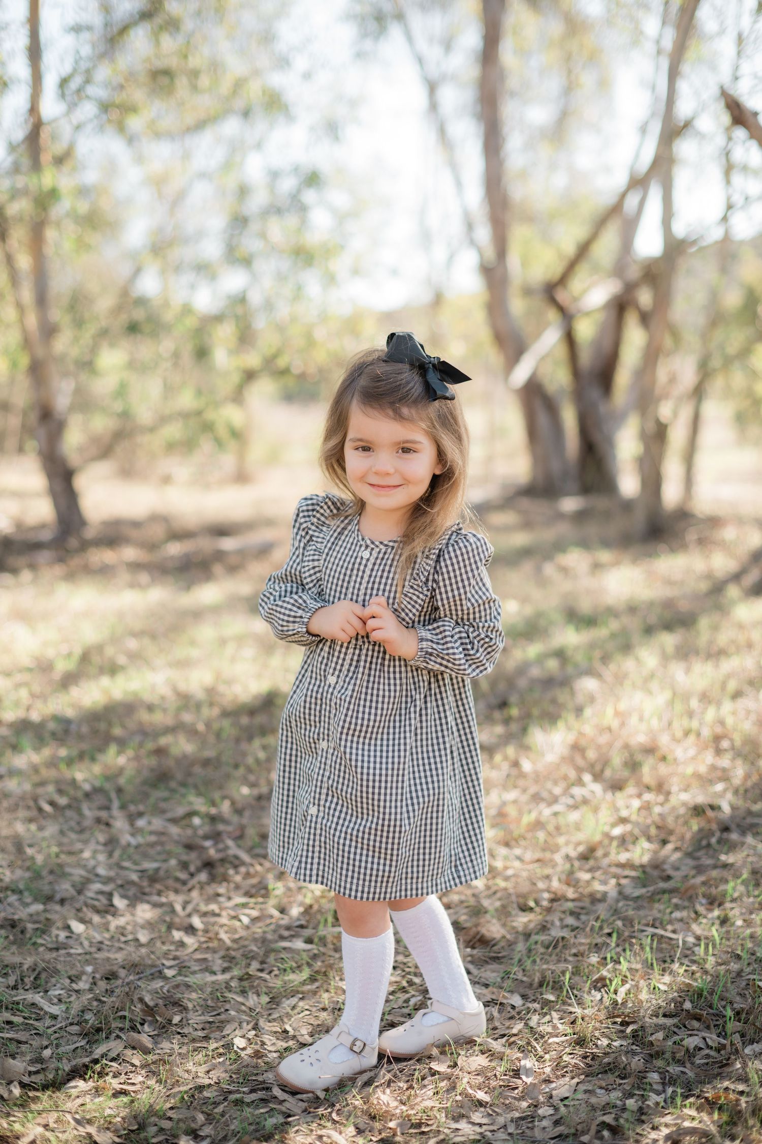 A smiling little girl in a plaid dress and black bow poses outdoors in a sunlit wooded area.