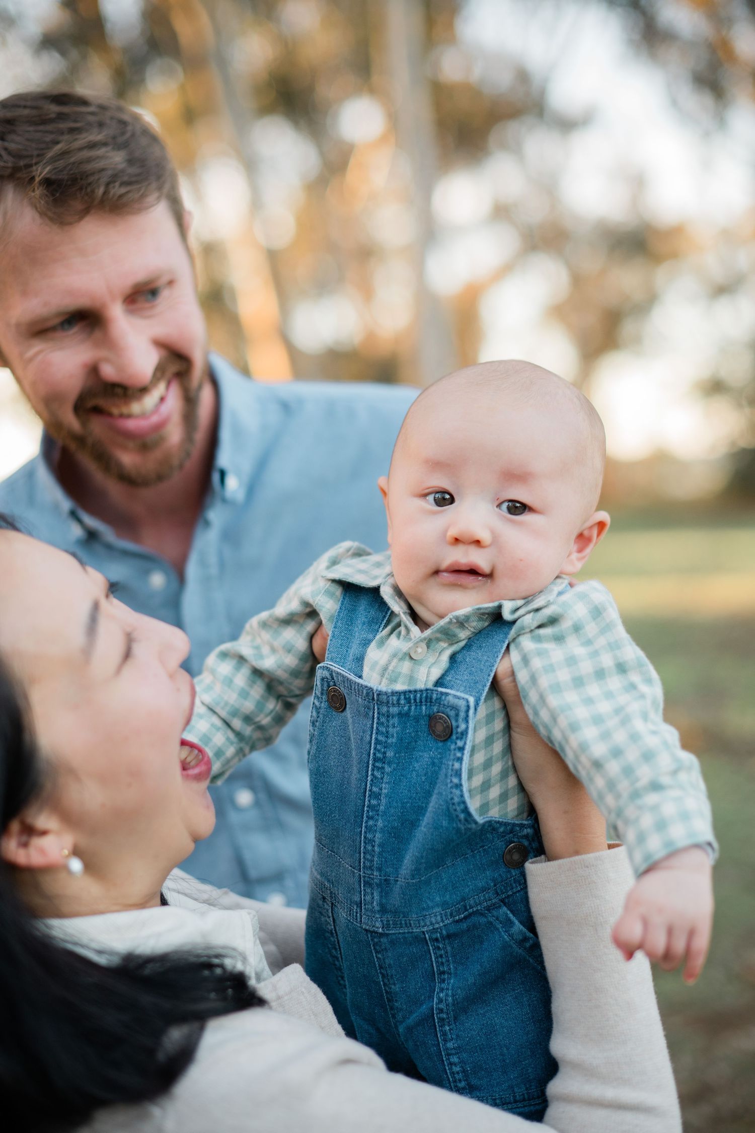 Happy family outdoors, parents smiling at adorable baby in denim overalls and plaid shirt, park setting.