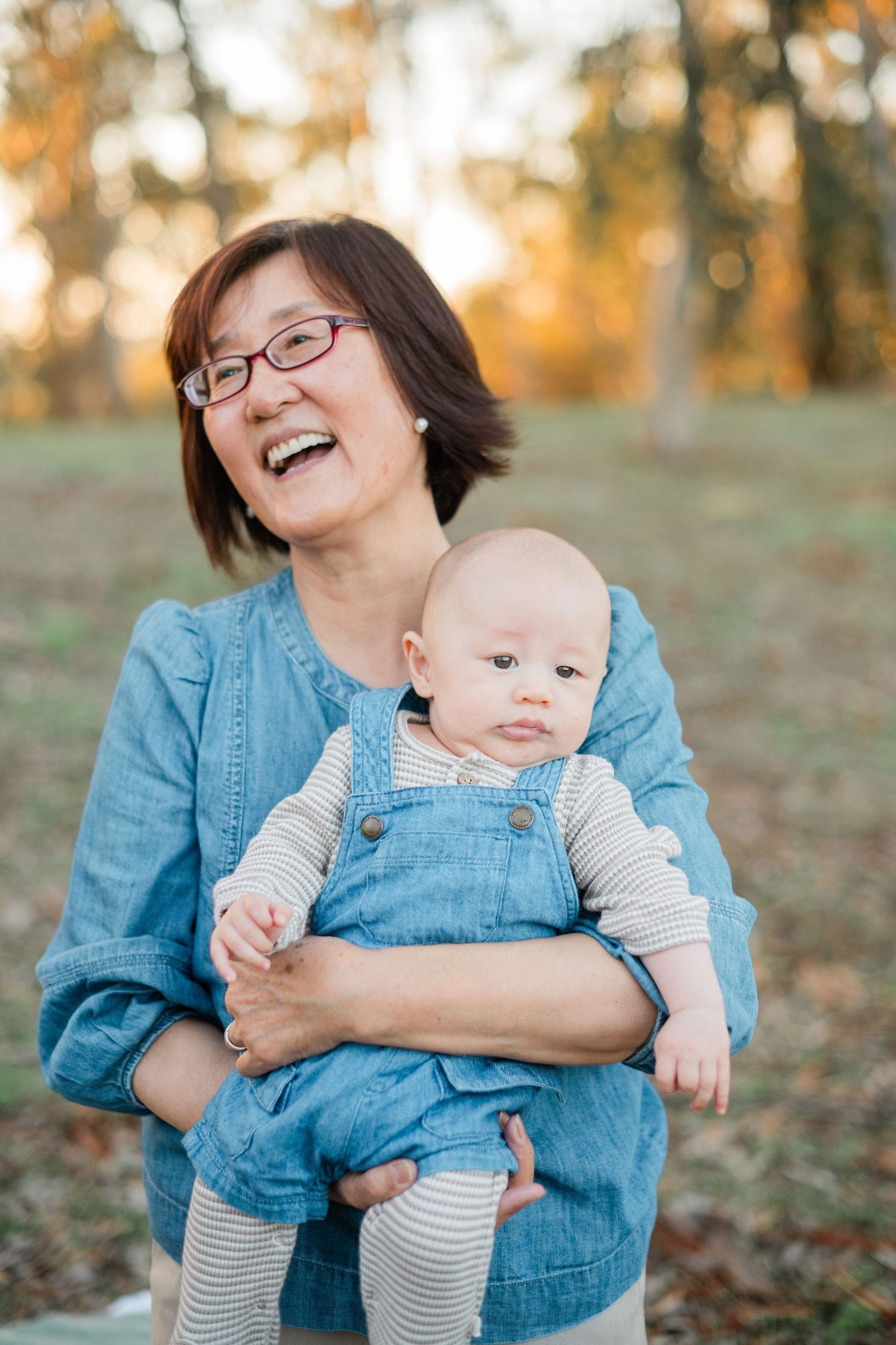 A smiling Asian grandmother wearing glasses holds a cute baby in denim overalls in a sunny autumn park.