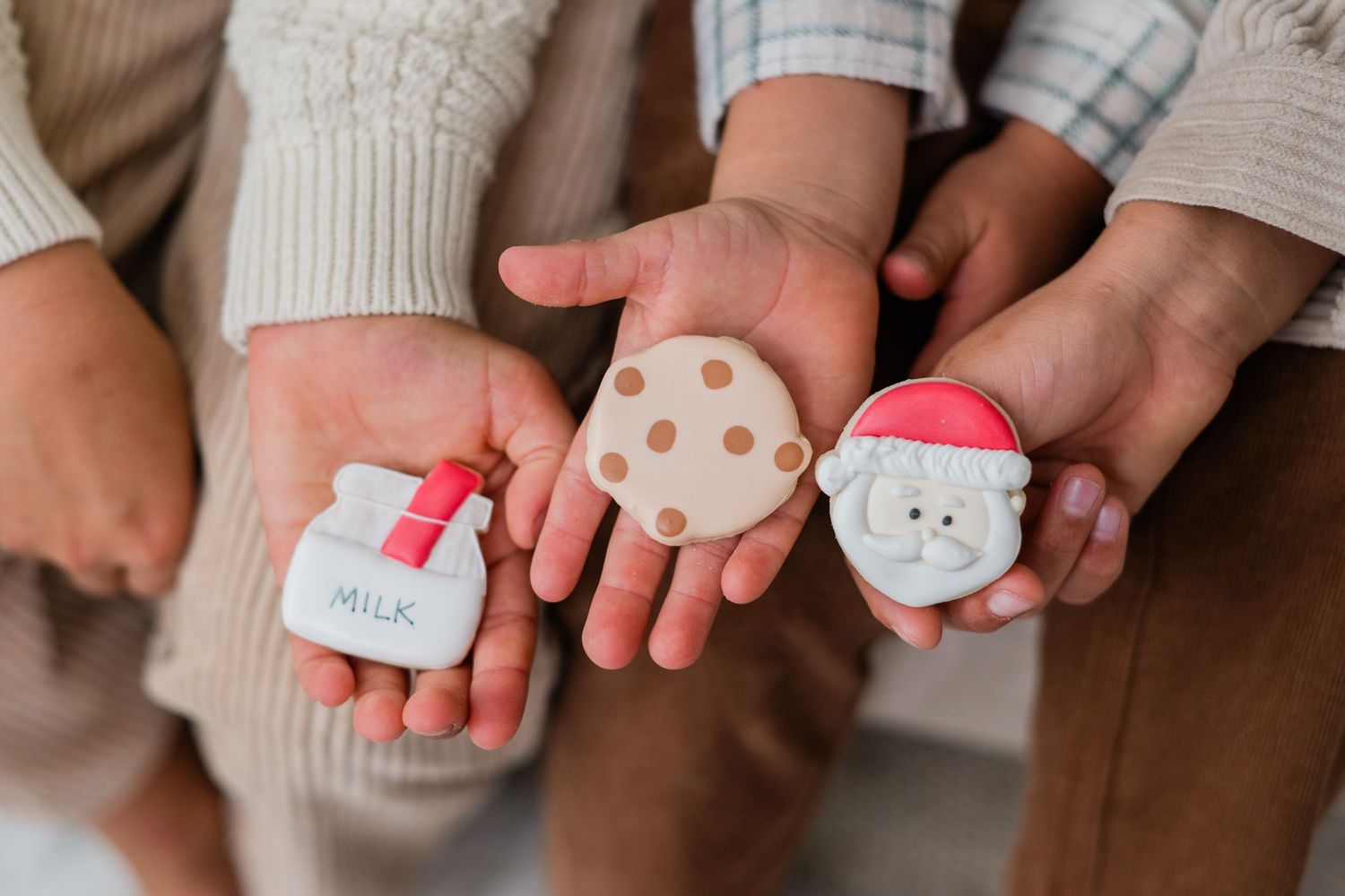 Three children hold out their palms displaying small Santa, cookie, and milk jug decorated Christmas sugar cookies.