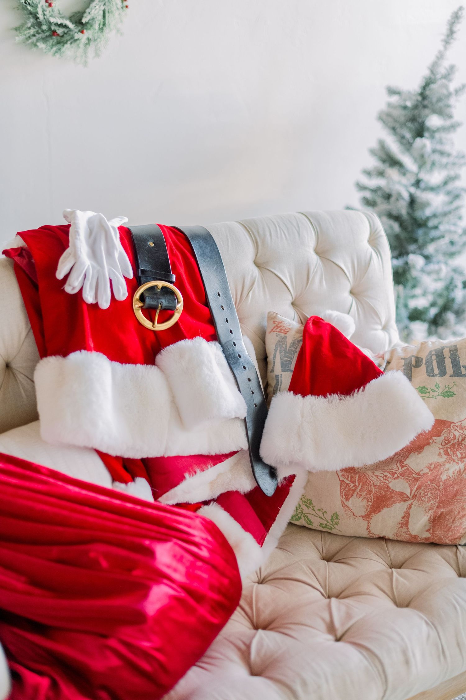 Santa Claus costume with red suit, white gloves, black belt, and hat laid on a cream tufted sofa near a Christmas tree.