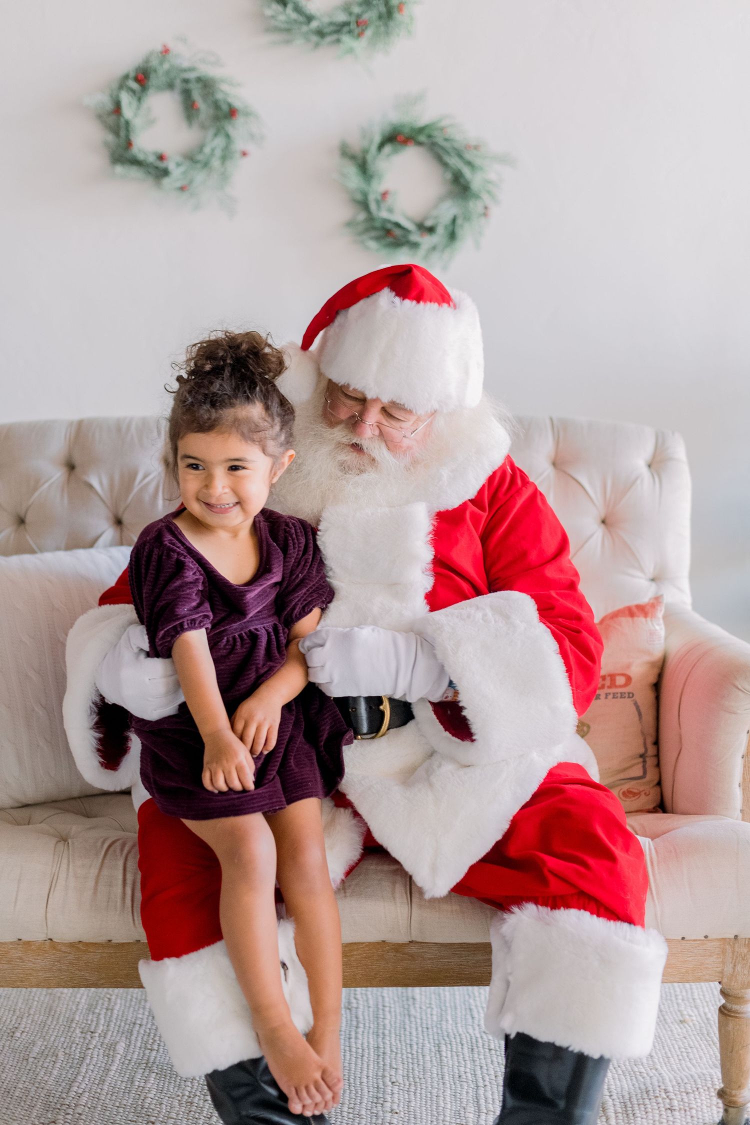 A young girl in a purple dress smiles while sitting beside Santa Claus on a beige couch with holiday wreaths.