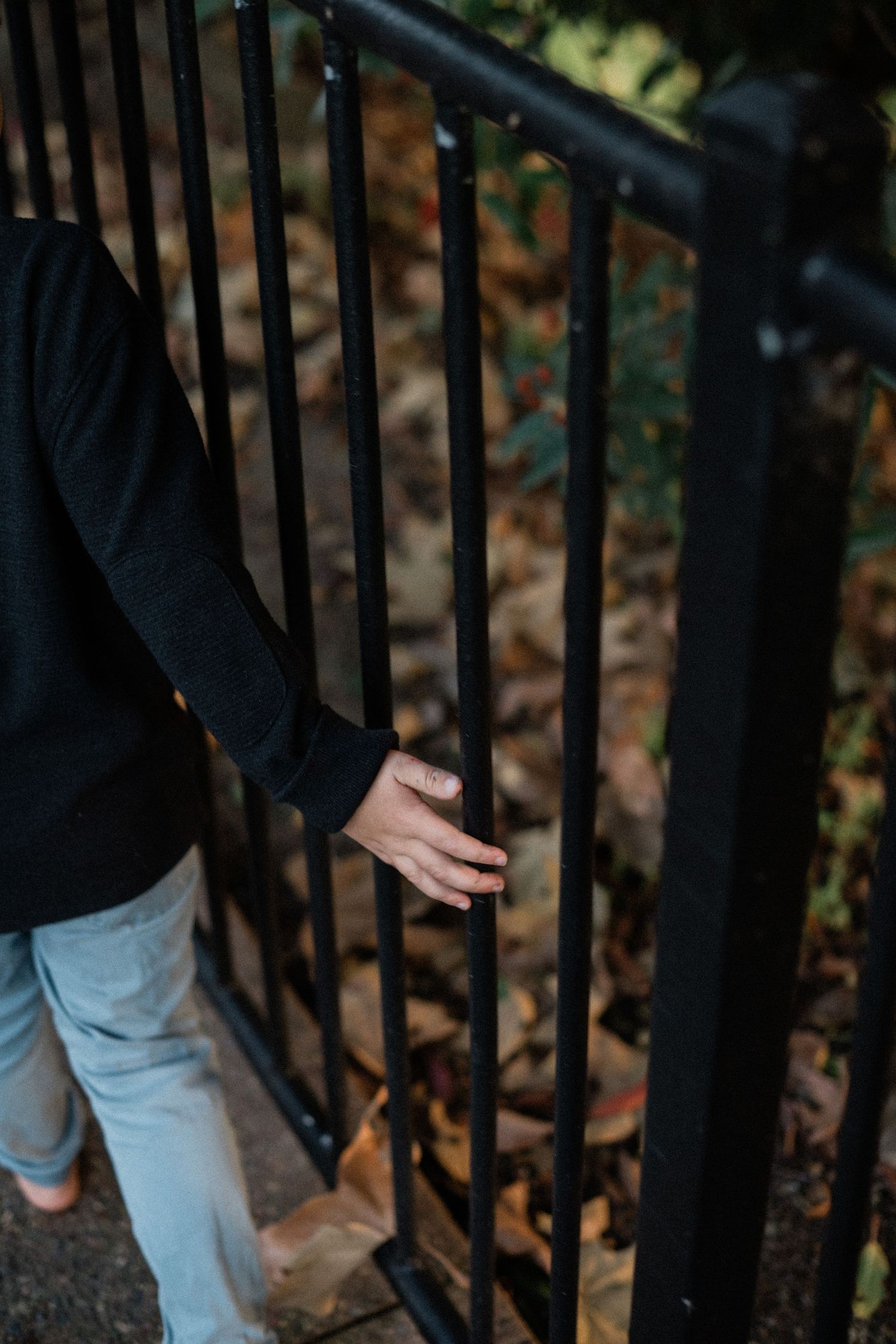 A child in a black sweater holds a black metal fence railing surrounded by autumn leaves.