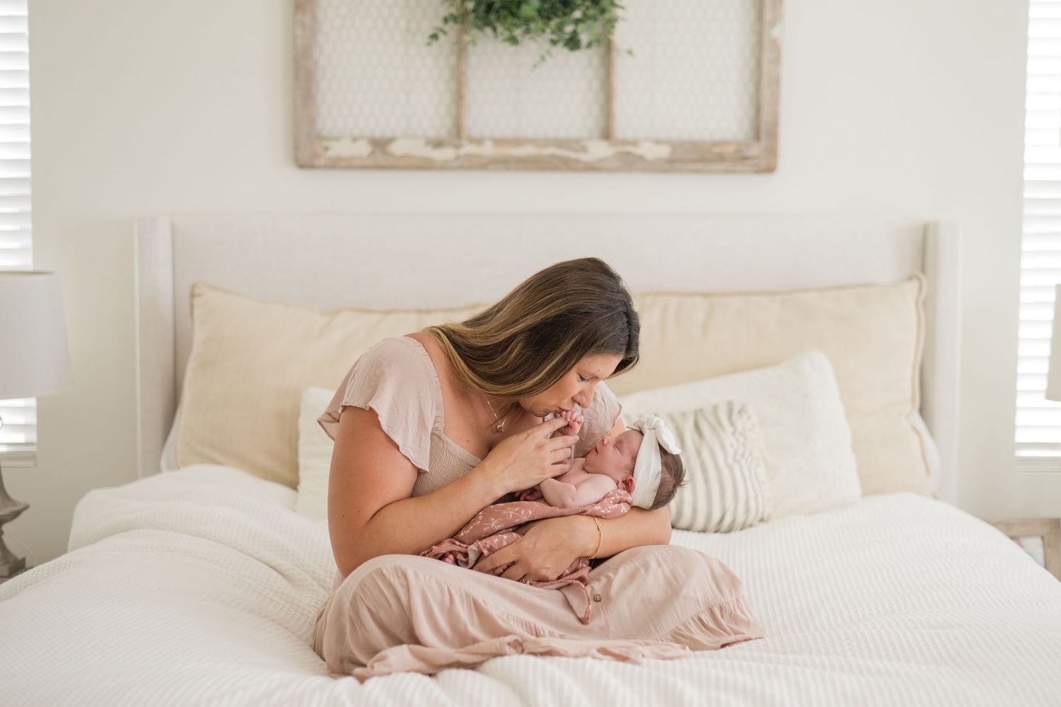 Mother tenderly kisses her newborn baby while sitting on a white bed in a bright, serene nursery bedroom.