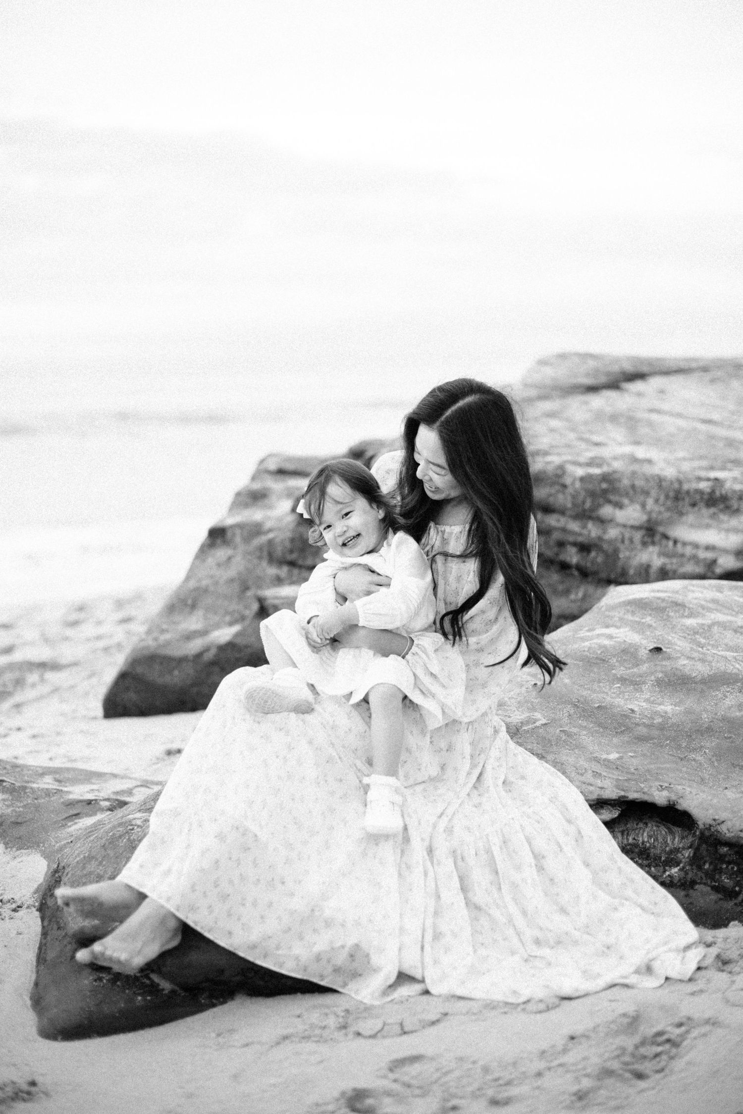 Black and white photo of a mother sitting on beach rocks, lovingly holding her young daughter in a floral dress.