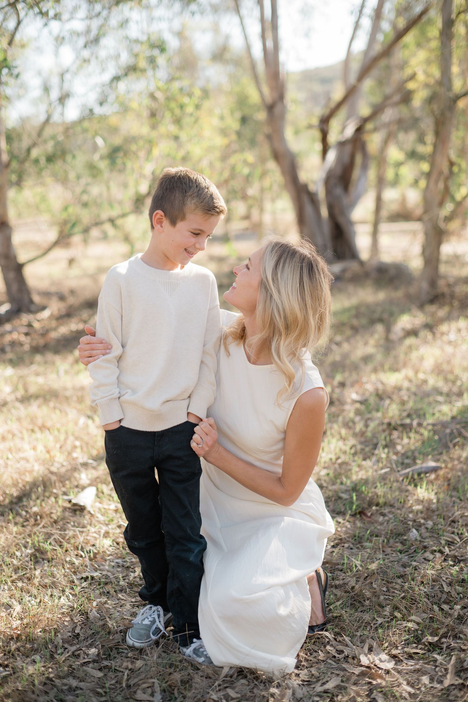 A blonde woman kneels beside her son, sharing a warm smile in a sunny outdoor setting.