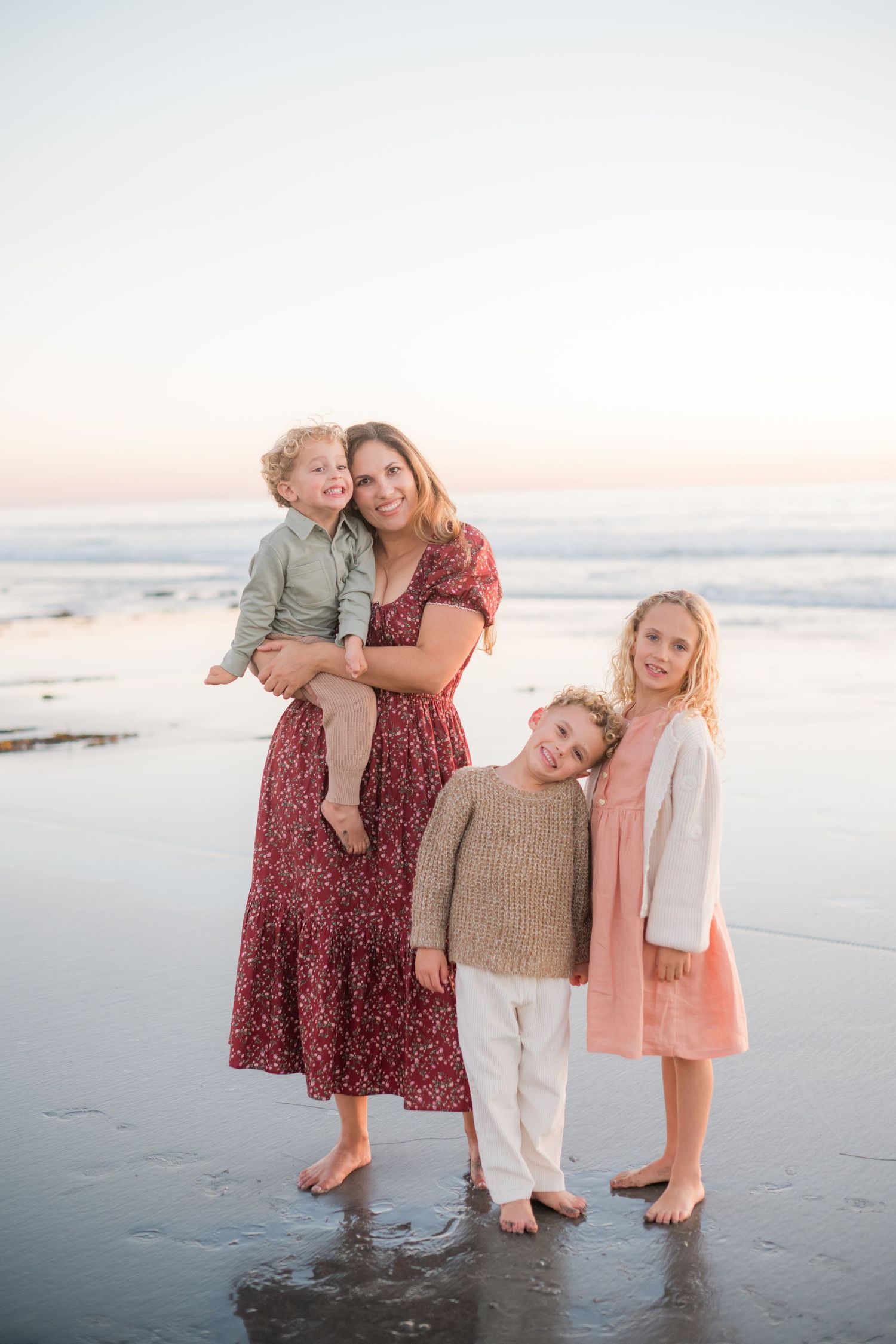 Mother in red floral dress holds toddler while two children lean on her at sunset beach.