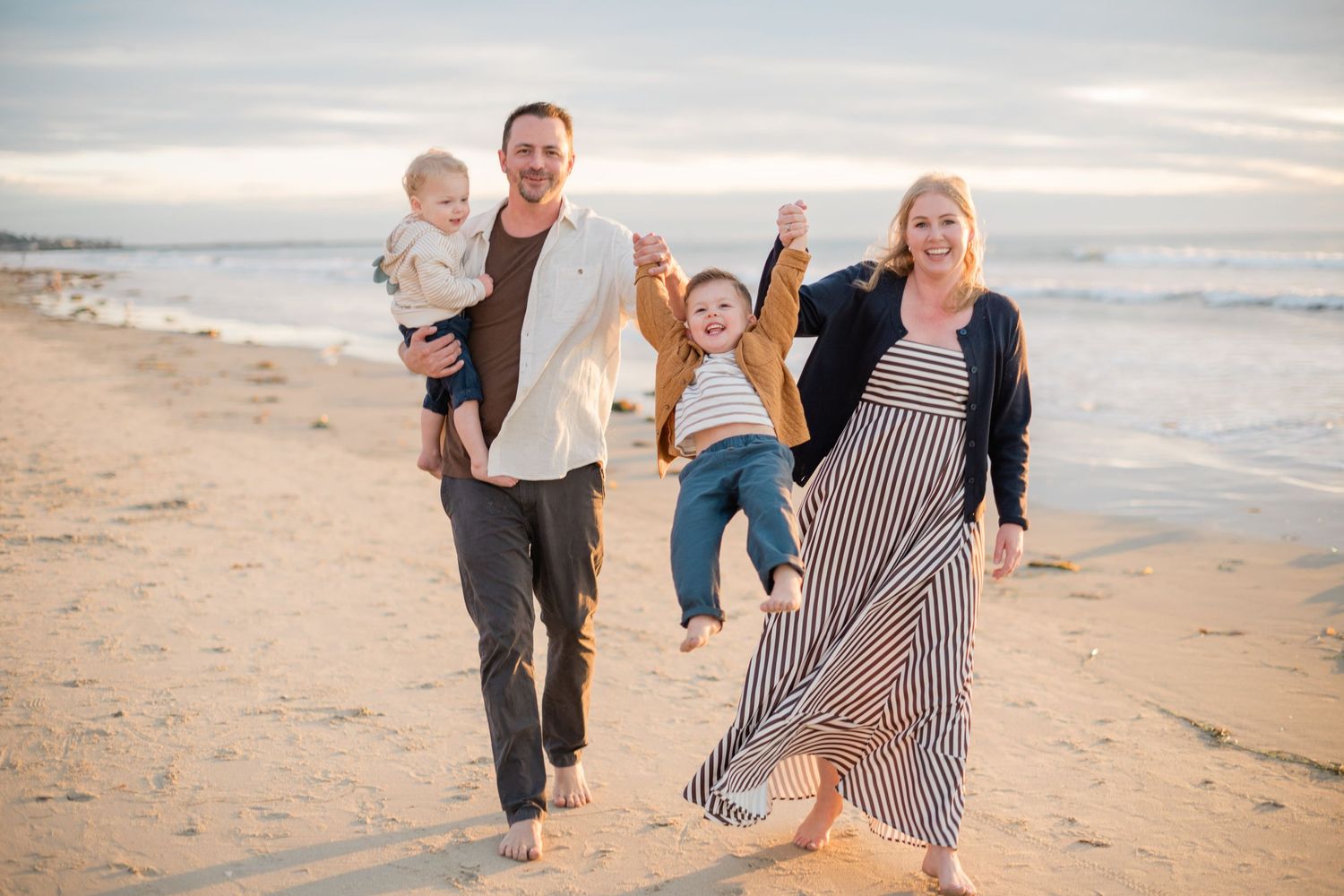 Happy family of four walking on a sandy beach at sunset, parents swinging children between them.
