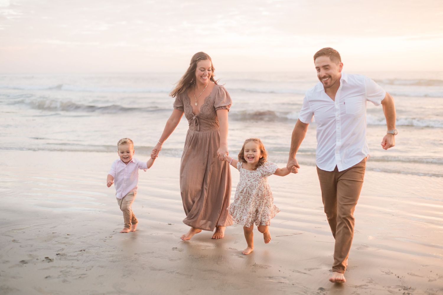 Family of four walking on beach at sunset, parents holding hands with two young children near ocean waves.