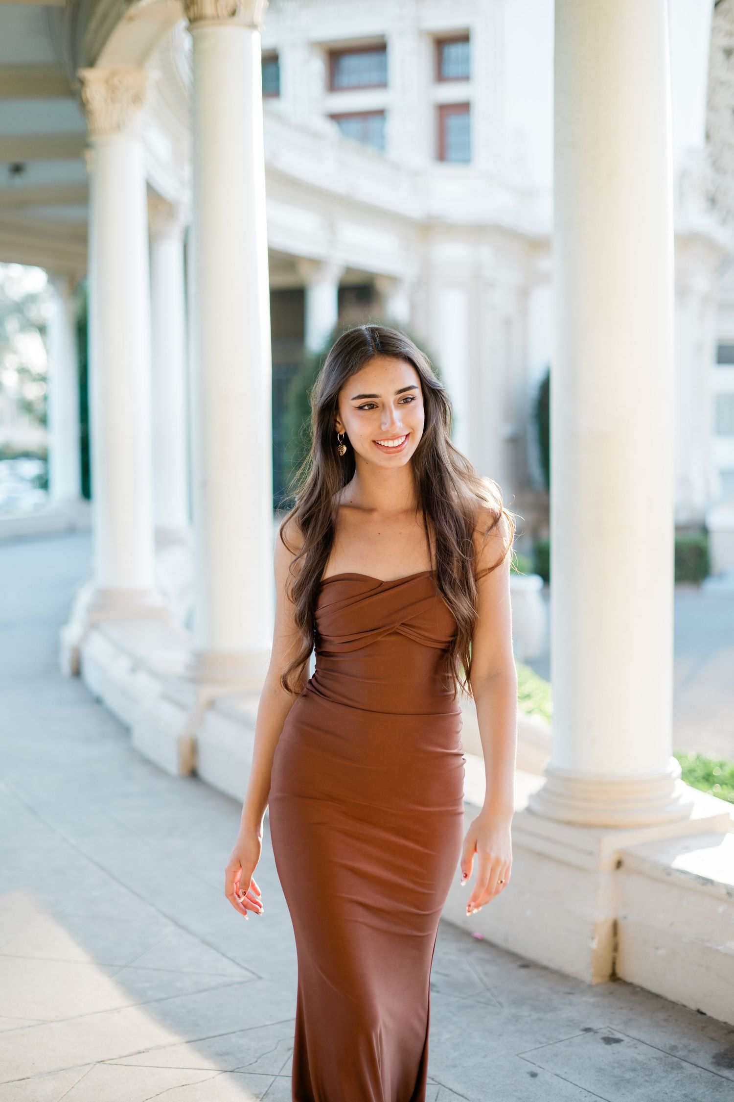 Young woman in brown off-shoulder maxi dress smiling among white columns.