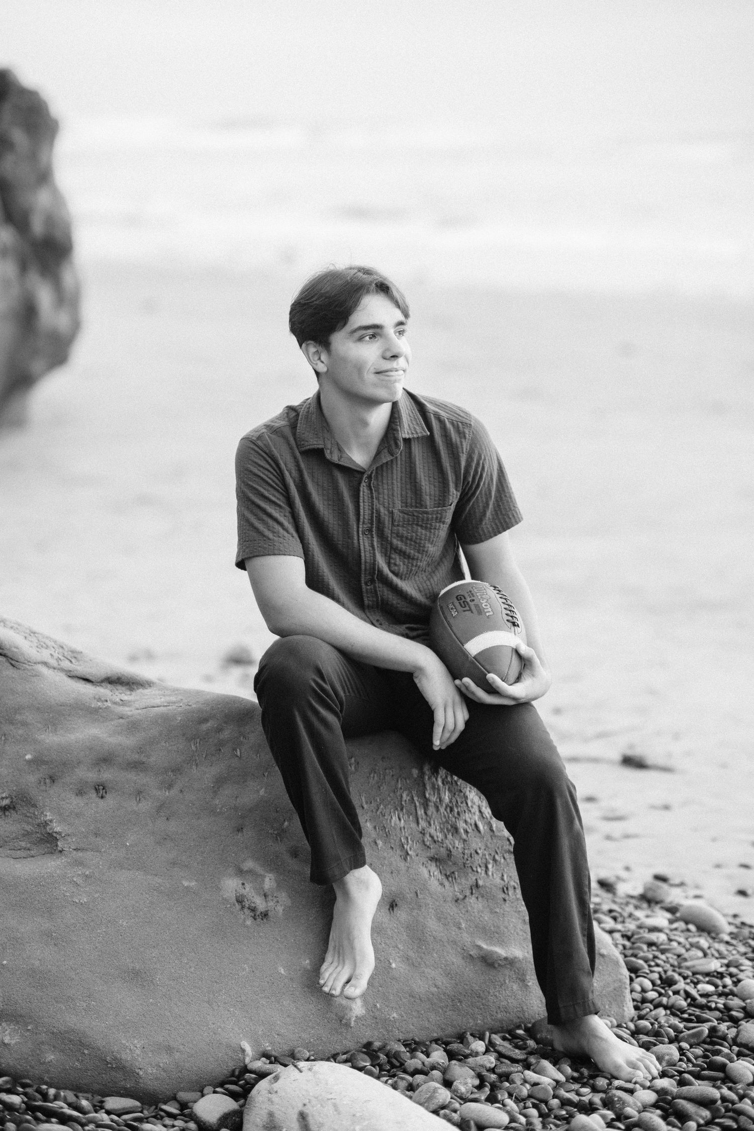 Young man sitting barefoot on a rock at the beach holding a football, black and white portrait photo.