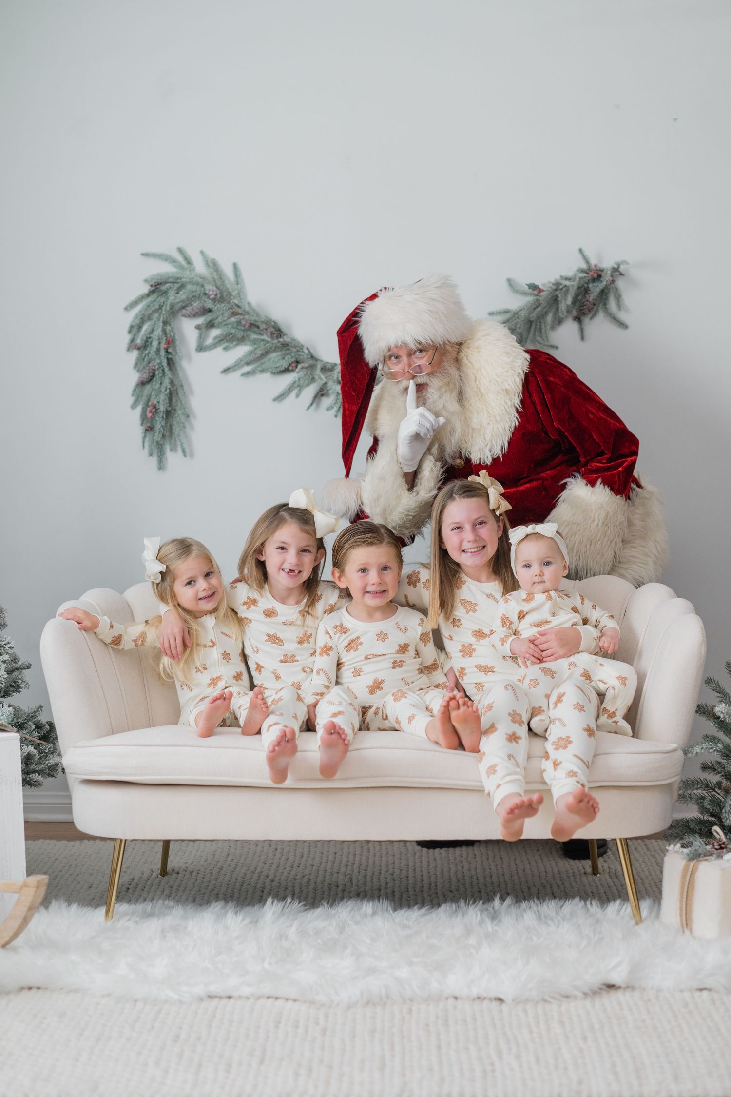 Five young girls in matching pajamas sit on a white sofa with Santa Claus shushing behind them.