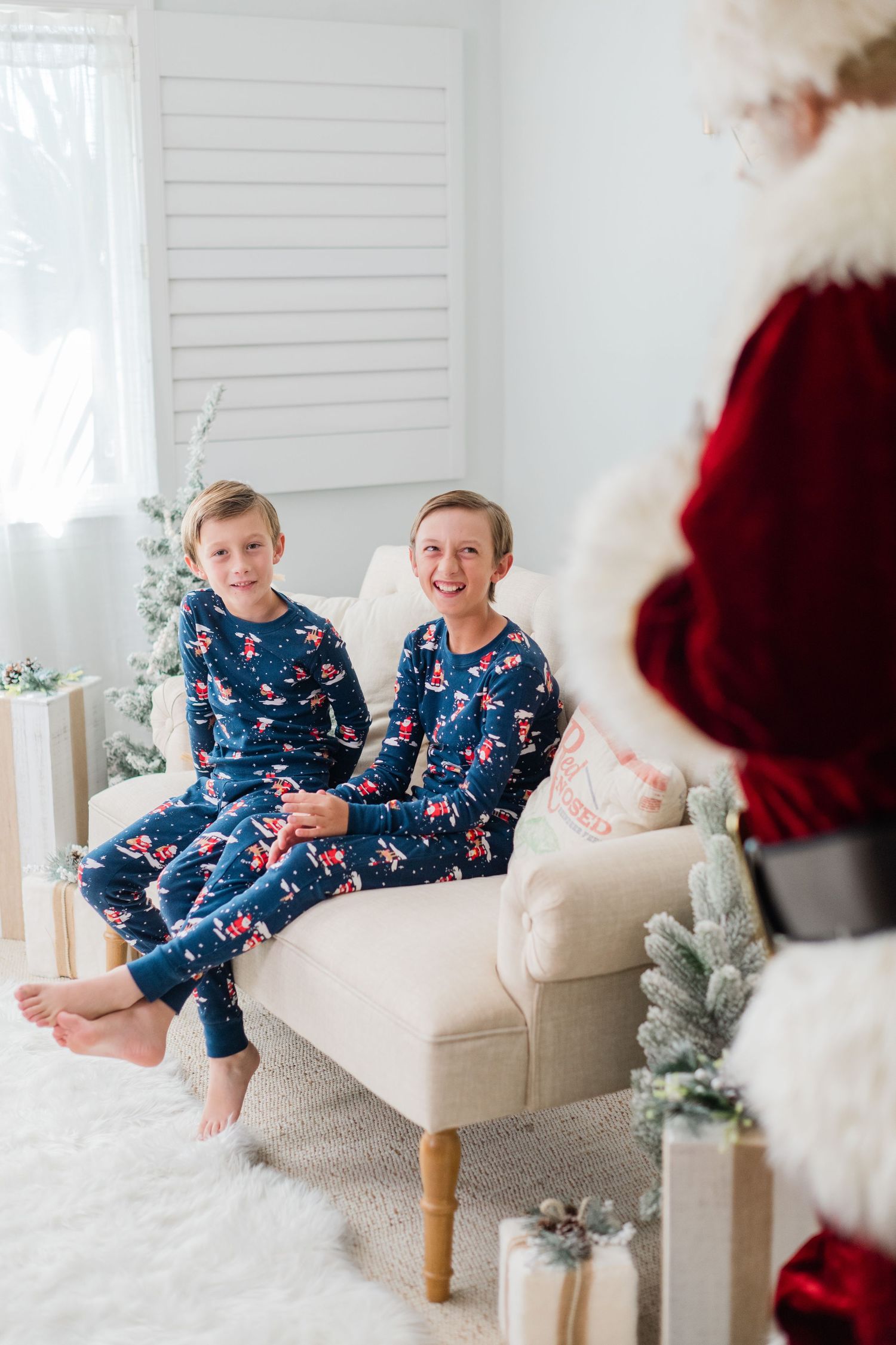 Two boys in matching blue Christmas pajamas sitting on a chair smiling at Santa Claus during a holiday photo session.
