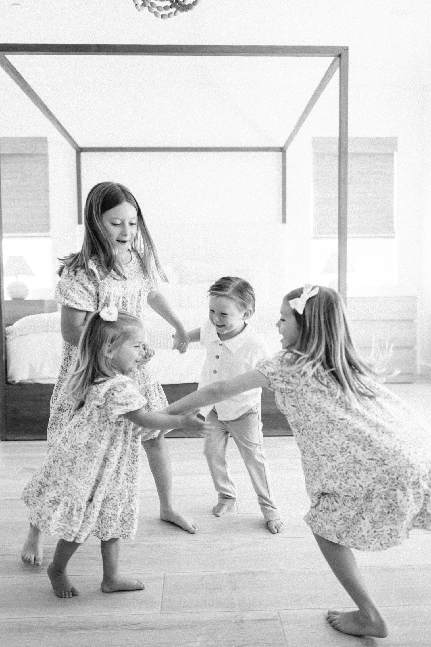 Black and white photo of a mother and three young children playing and dancing together in a bright bedroom.