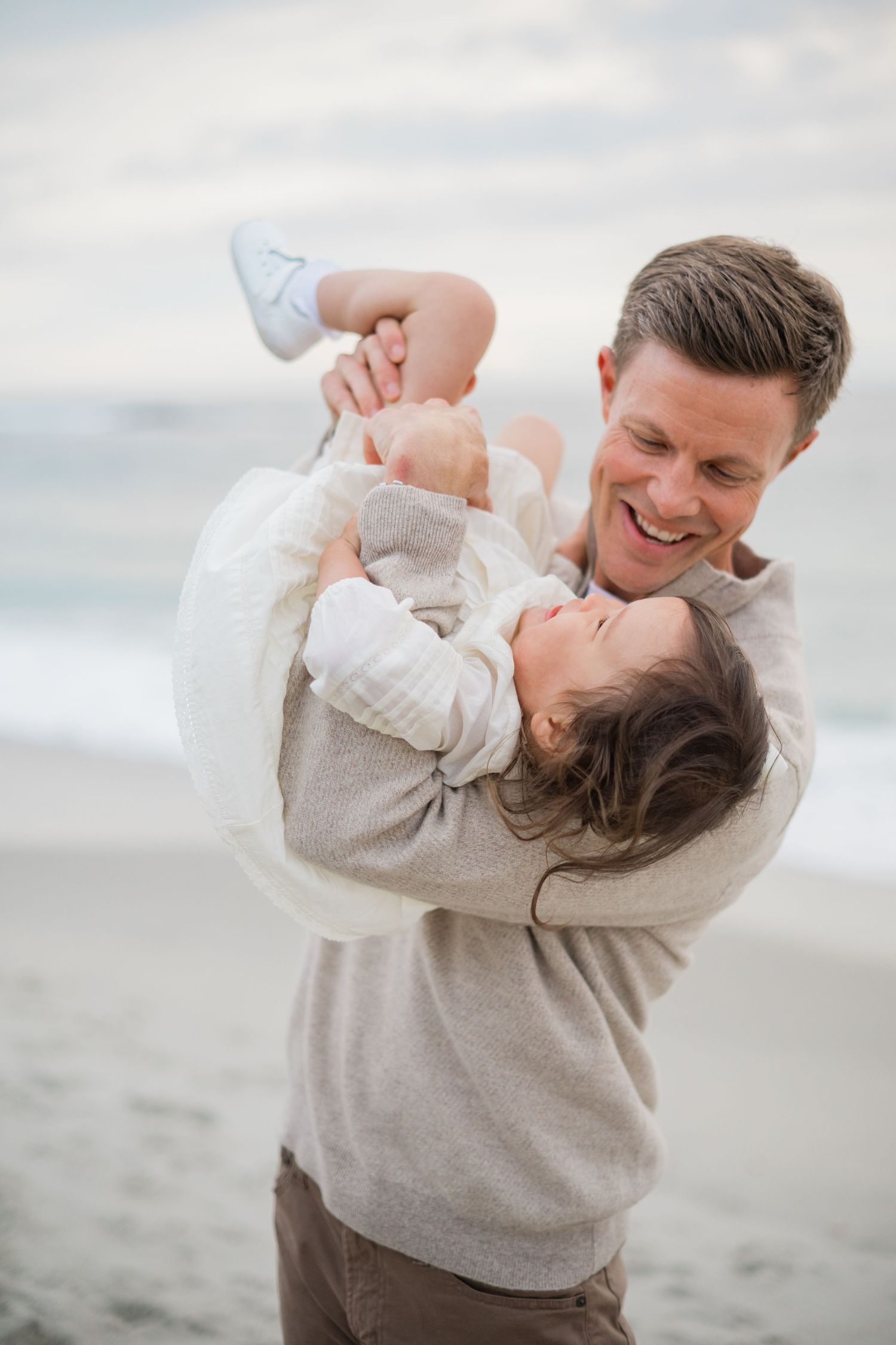 A smiling father joyfully lifts his baby at the beach, both wearing neutral tones against an ocean backdrop.