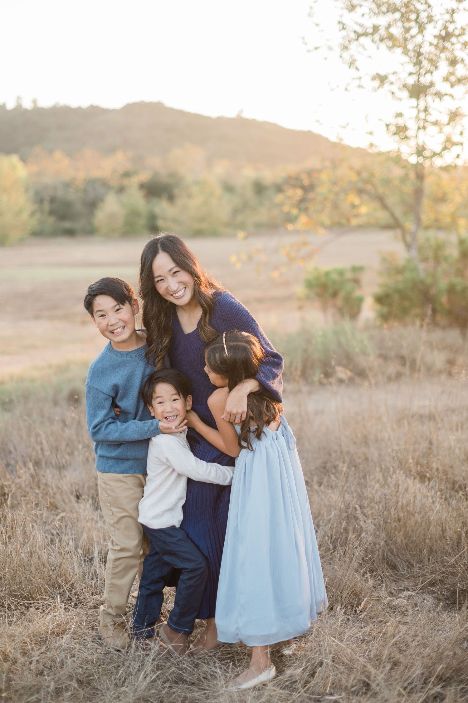 A smiling mother hugs her three young children in a sunlit field with golden hills and autumn trees in the background.