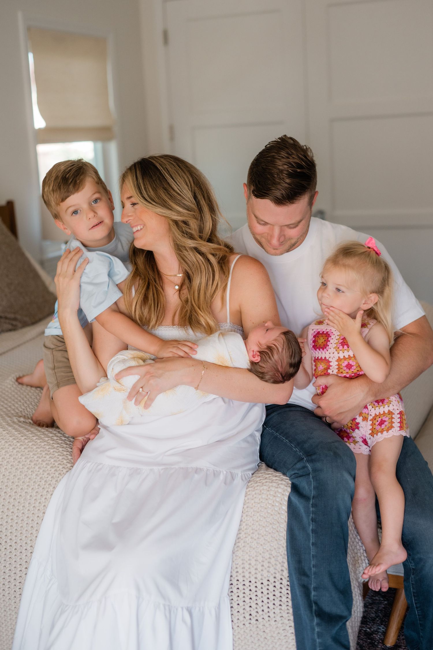Happy family of five sitting on a bed, parents holding three young children including a newborn baby.