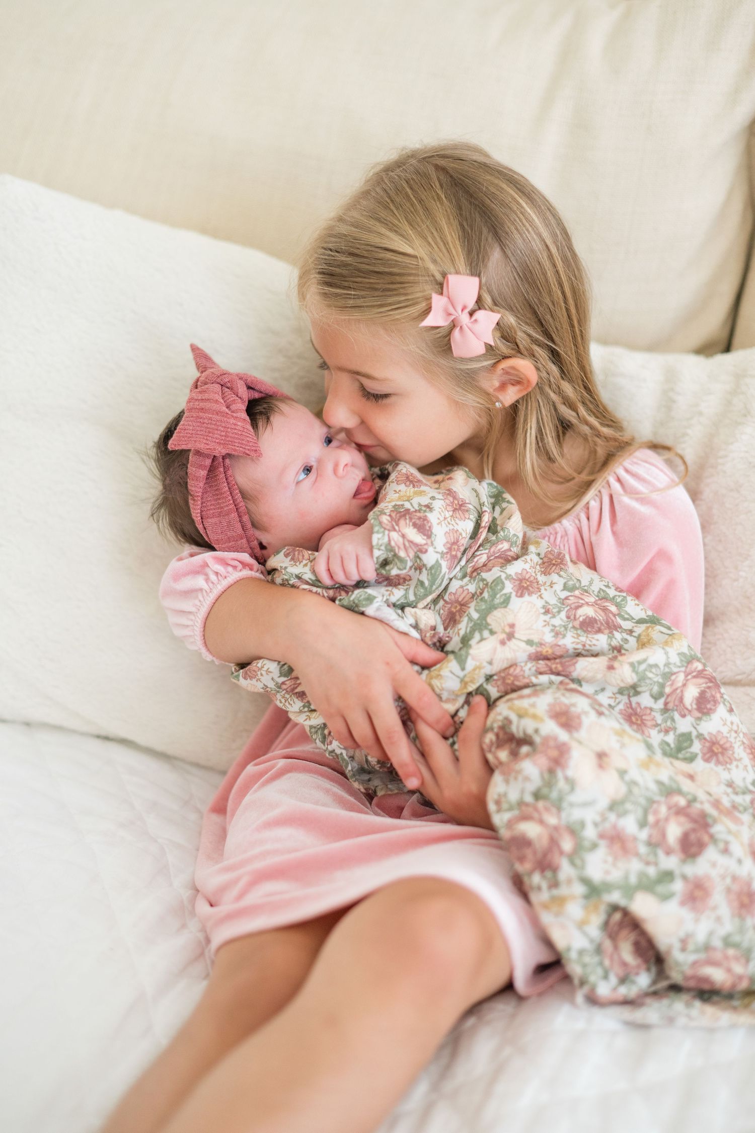 A young girl in pink dress kisses her newborn sibling wearing a floral outfit and pink headband on a white sofa.