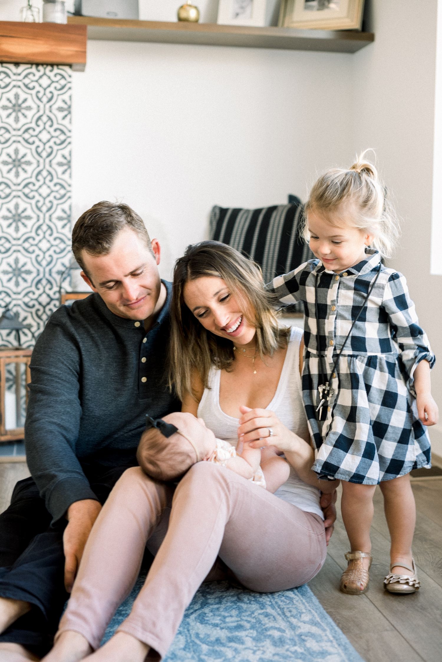 Happy family of four sitting on floor, parents smiling at newborn baby while toddler girl stands beside them.