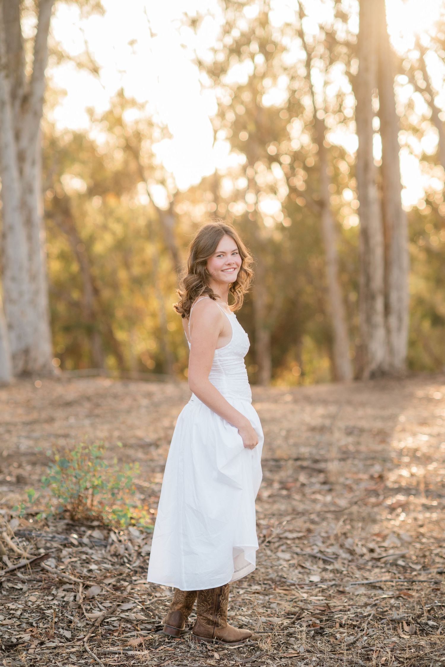 Young woman in white dress and cowboy boots smiling in golden hour forest setting.